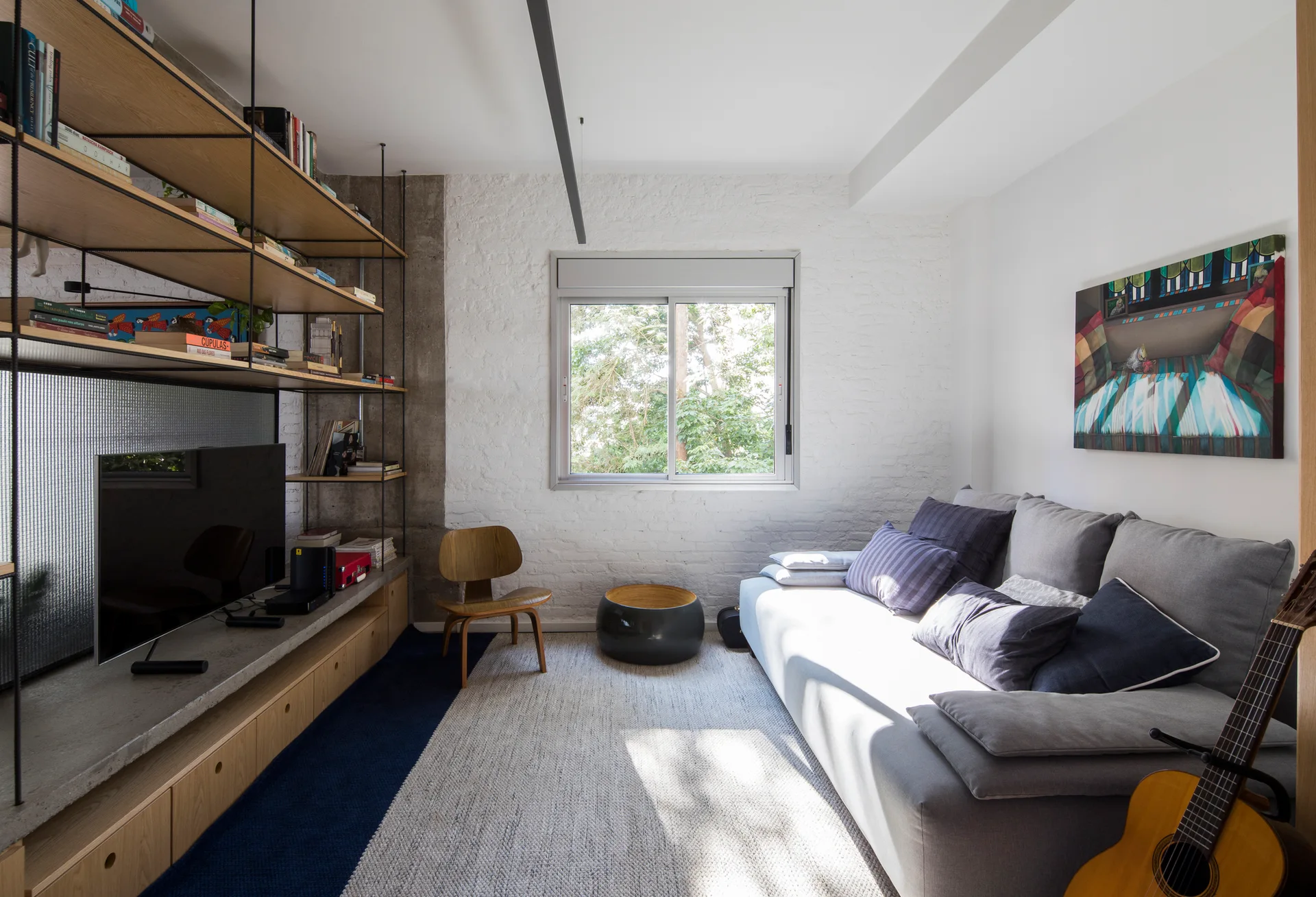 A modern living room featuring a grey fabric sofa, an open wood and metal shelving unit, and a window set against a white painted brick wall.
