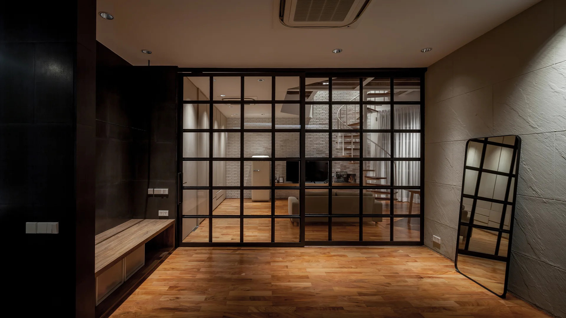 A modern interior features a black metal grid glass partition separating a wood-floored entryway from a living area with a spiral staircase.