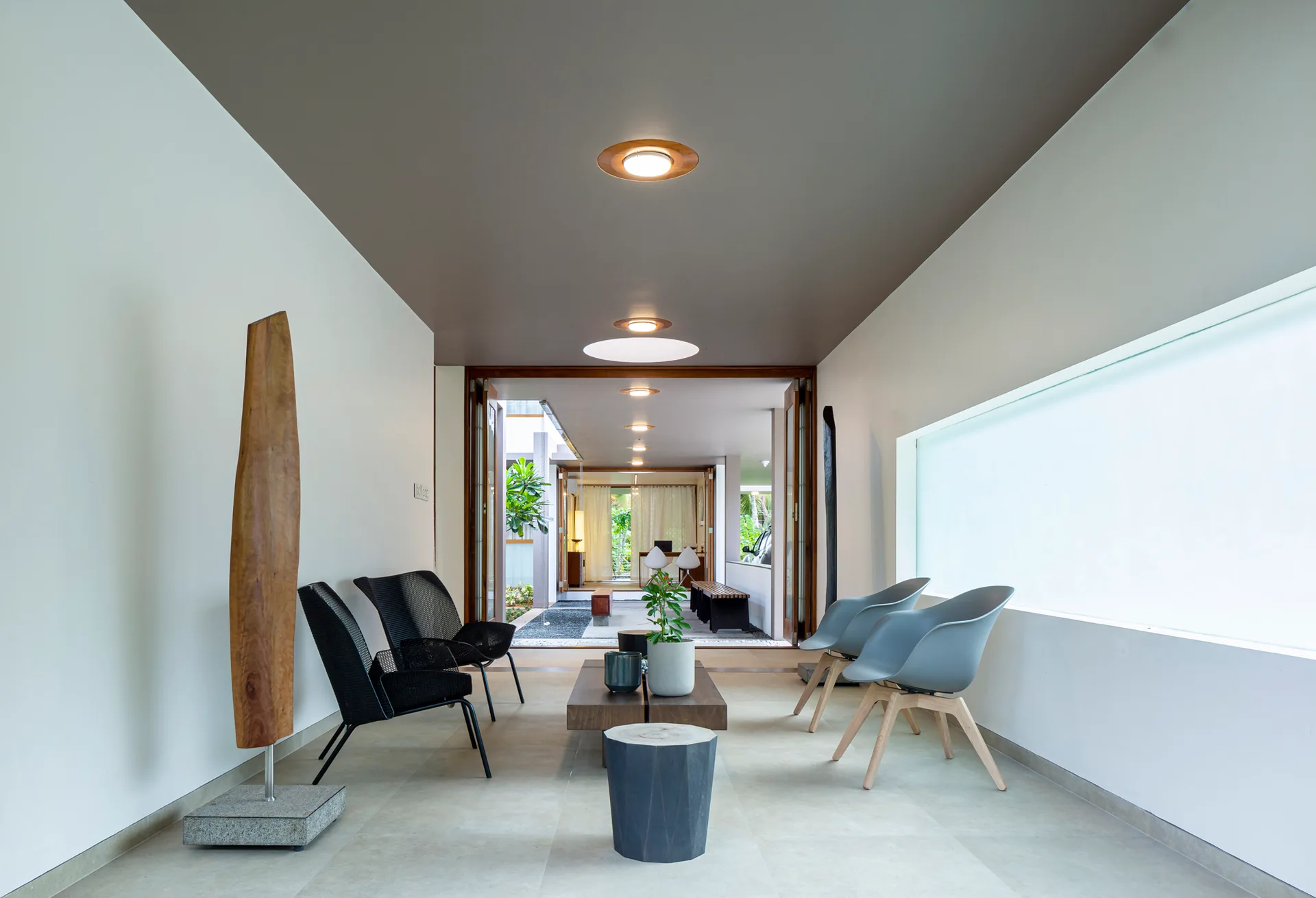 A minimalist lounge features asymmetrical black and gray chairs, a low wood coffee table, and a gray painted ceiling looking out toward an open courtyard.