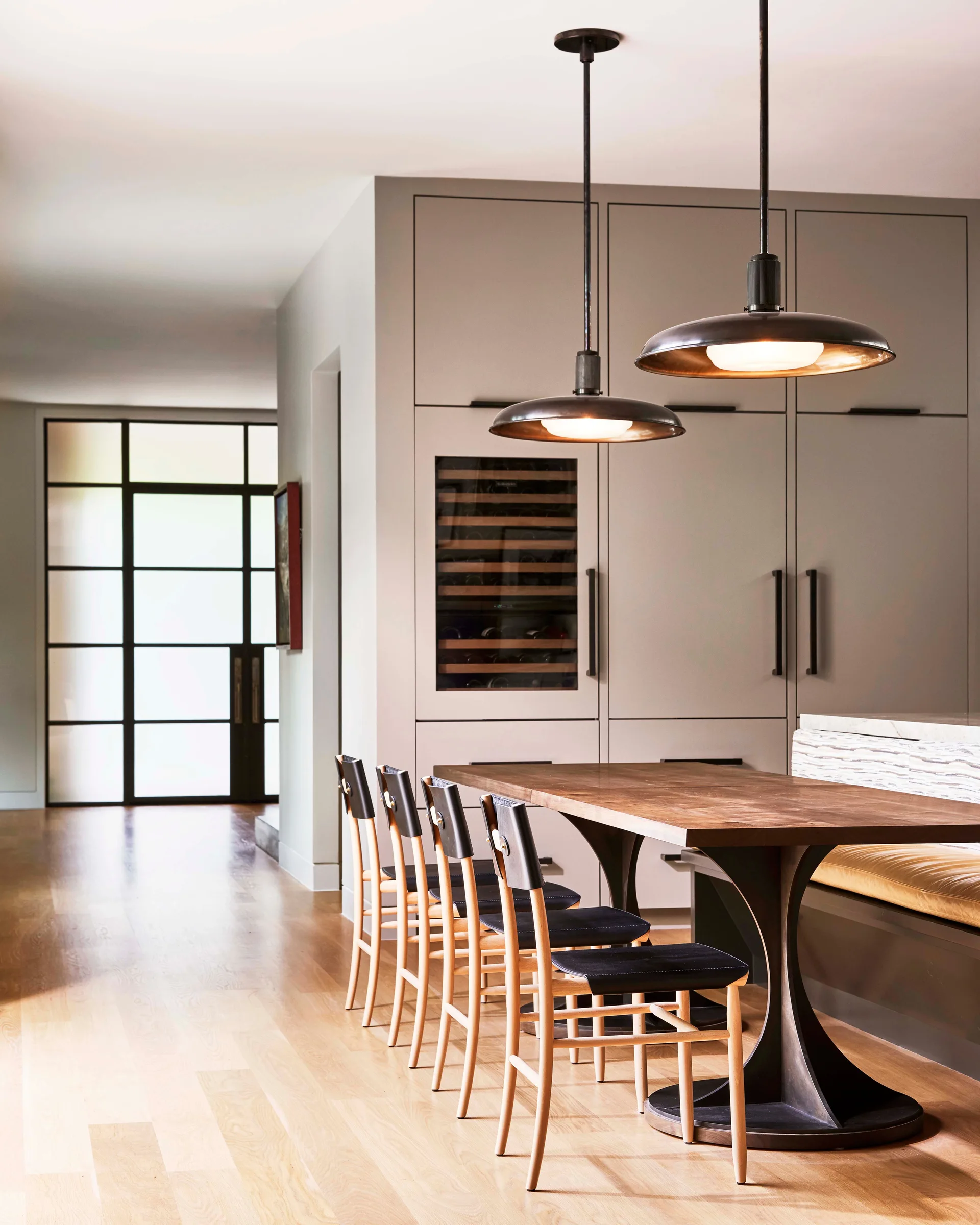 A wooden dining table with leather-seated chairs and a banquette sits in front of tall grey cabinets with an integrated wine fridge.