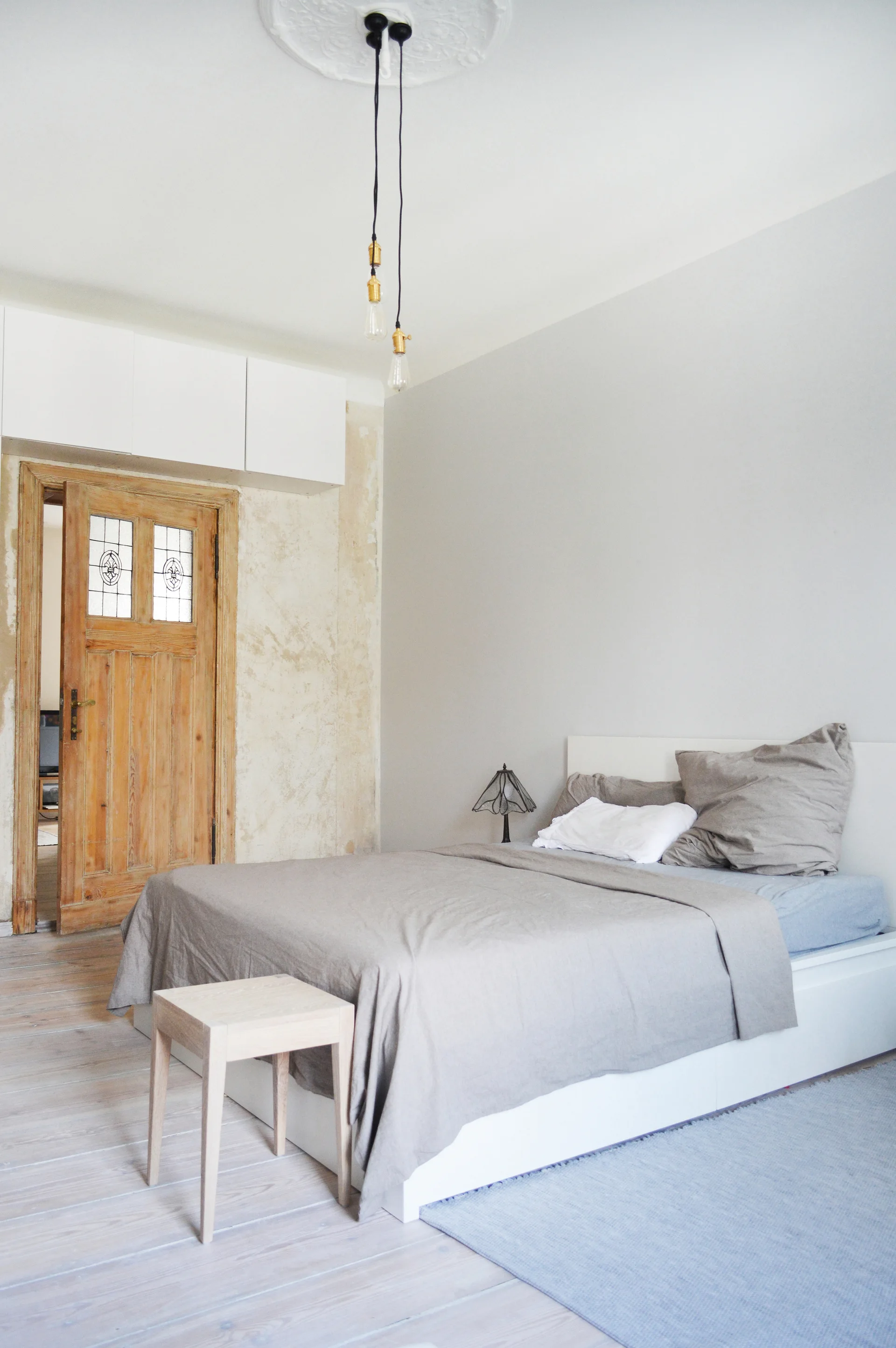 A minimalist bedroom features a bed with grey linens, a whitewashed wood floor, and a vintage wooden door under overhead cabinets.