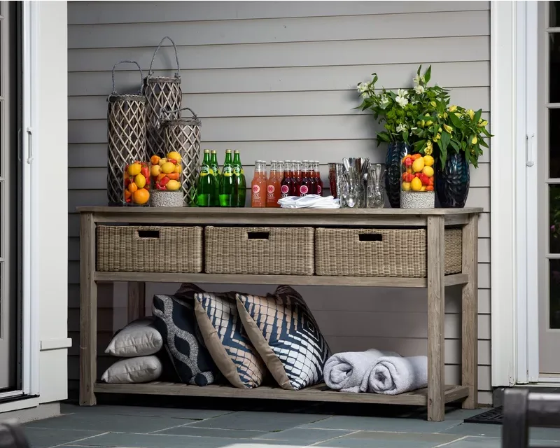 A weathered wood console table on a stone patio features wicker storage baskets, colorful beverages, decorative jars, and rolled towels.