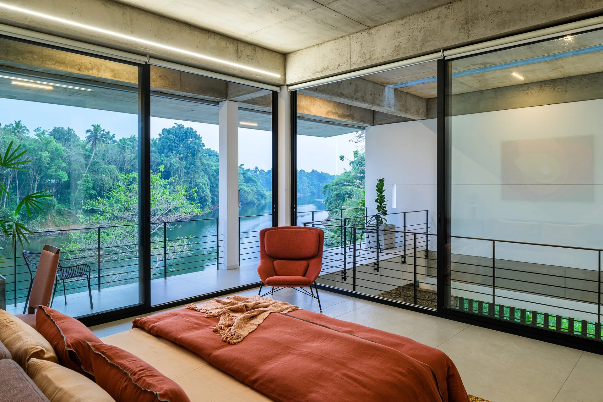 A modern bedroom features terracotta bedding, an orange armchair, an exposed concrete ceiling, and sliding glass doors overlooking a scenic river.