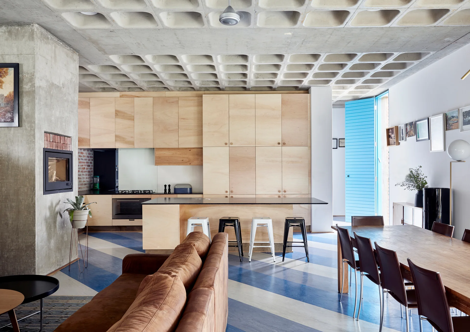 An open-concept kitchen and dining area features a concrete waffle ceiling, flat-front plywood cabinets, and a blue and white striped floor.