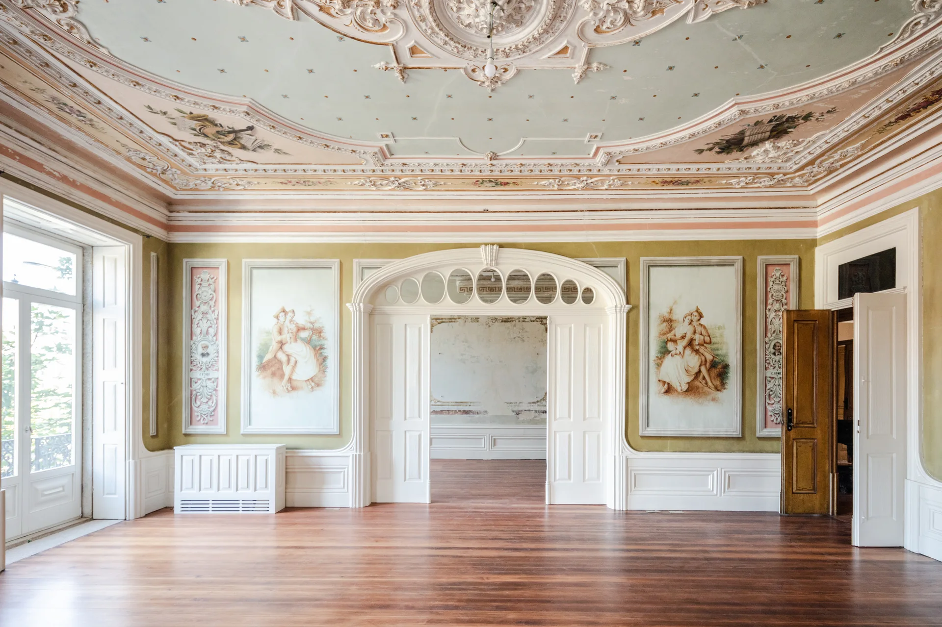 An empty historic room featuring polished wood floors, white wainscoting, wall murals of couples, and an ornate plaster ceiling.