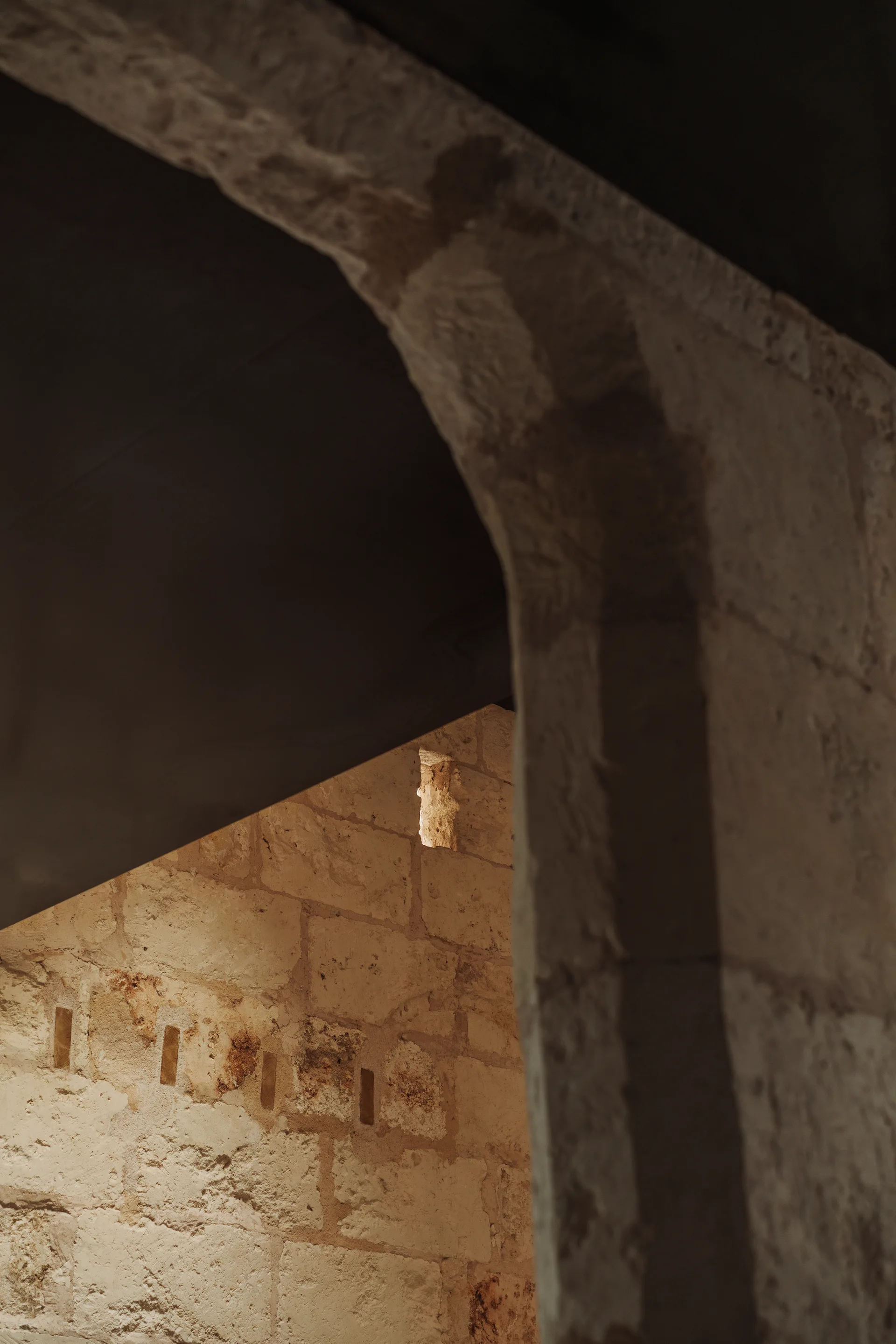 A rustic stone archway frames an exposed masonry wall illuminated by warm light in a dimly lit historic interior.