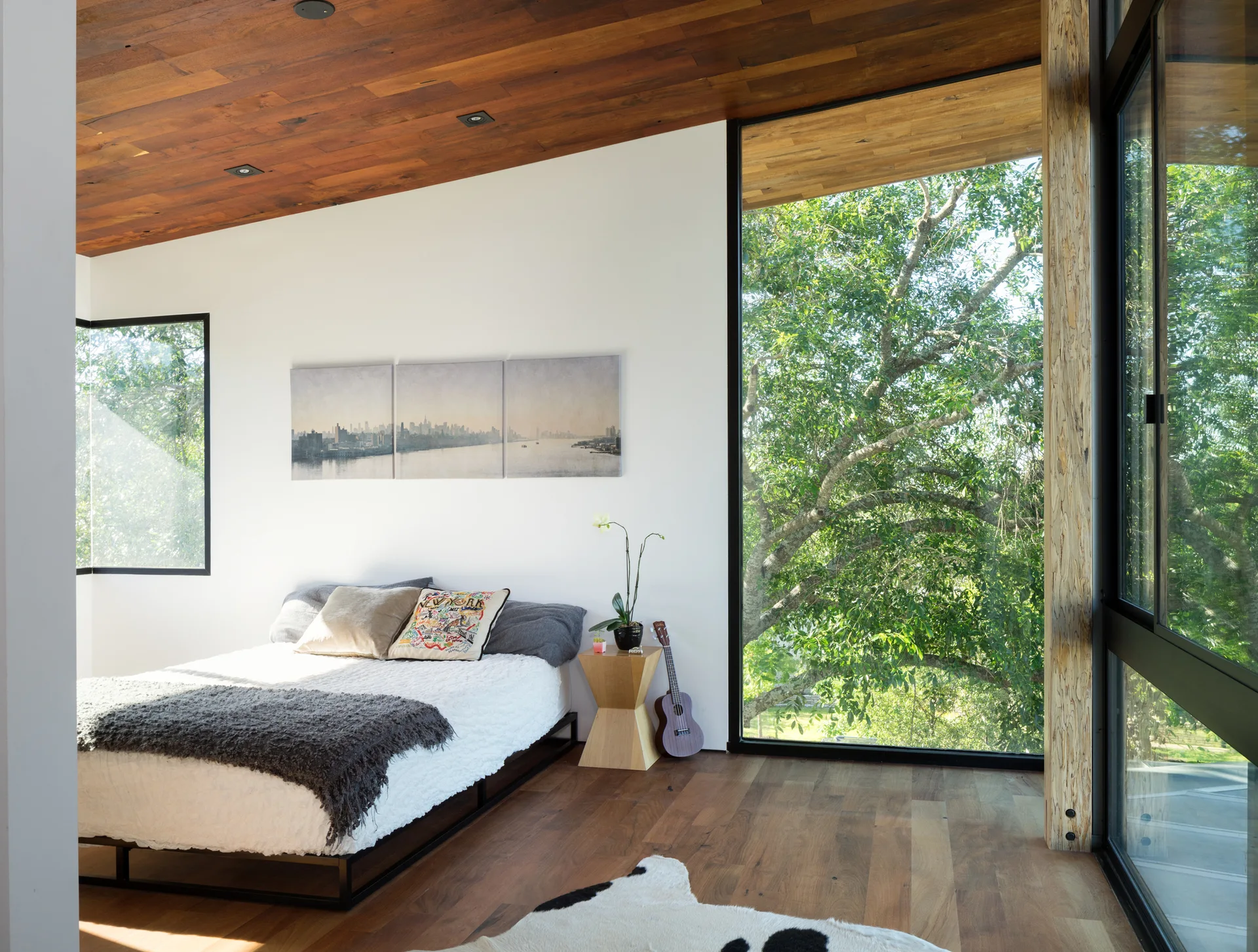 A modern bedroom features a platform bed, a geometric side table, and a wood plank ceiling next to large floor-to-ceiling windows overlooking trees.