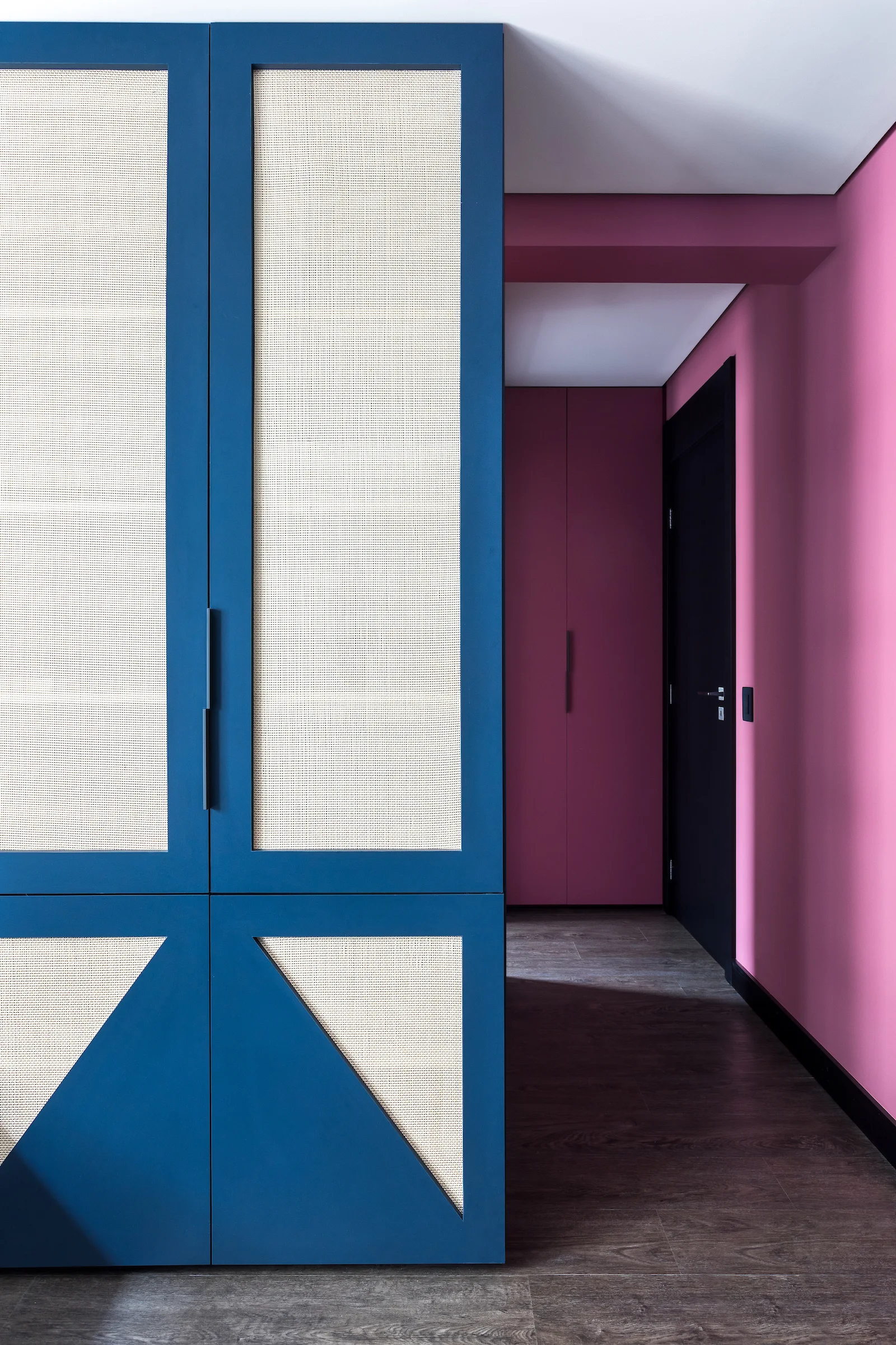 A custom blue wardrobe with woven mesh panels stands in a hallway featuring vibrant pink walls, a dark wood floor, and a black door.