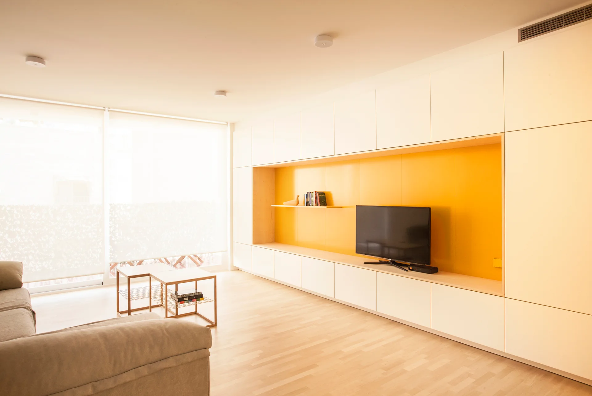 A bright living room features a white wall-to-wall media unit with a yellow recessed niche, a flat screen television, and a beige sofa.
