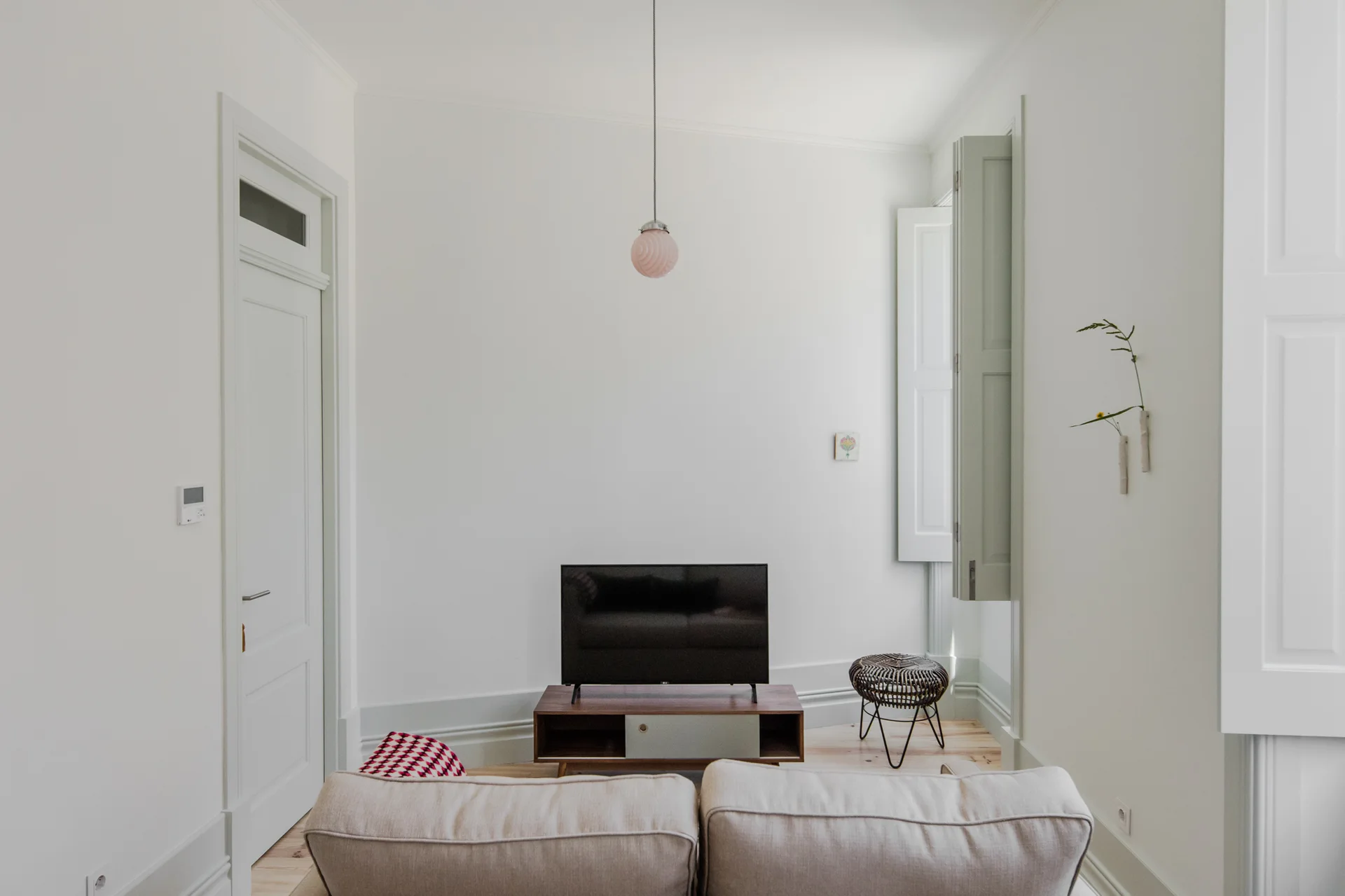 A minimalist white living room features a beige sofa in the foreground, a low wood media console with a television, and pale green window shutters.
