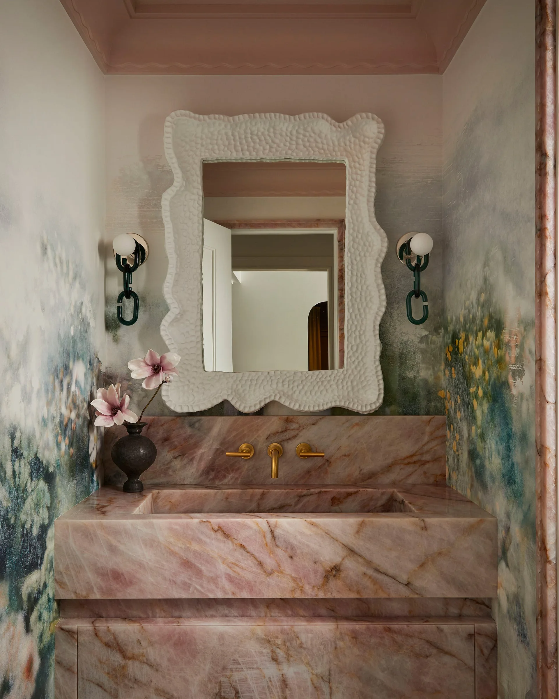A pink veined marble integrated vanity stands below a textured white mirror and green scenic wallpaper in a powder room.