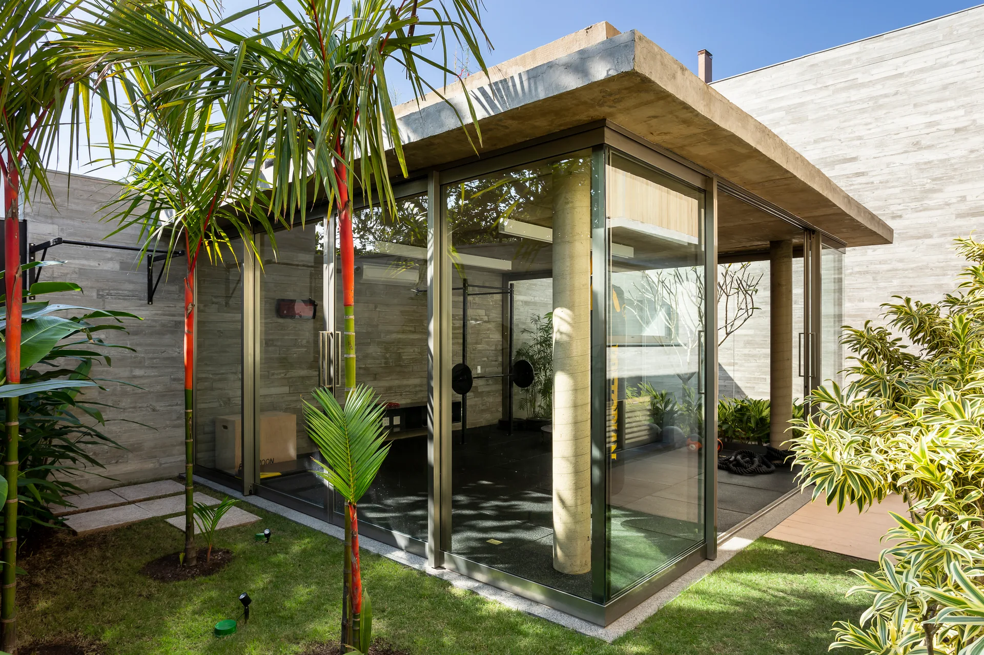A modern freestanding glass gym pavilion with a flat concrete roof is situated in a green backyard garden surrounded by palm trees.