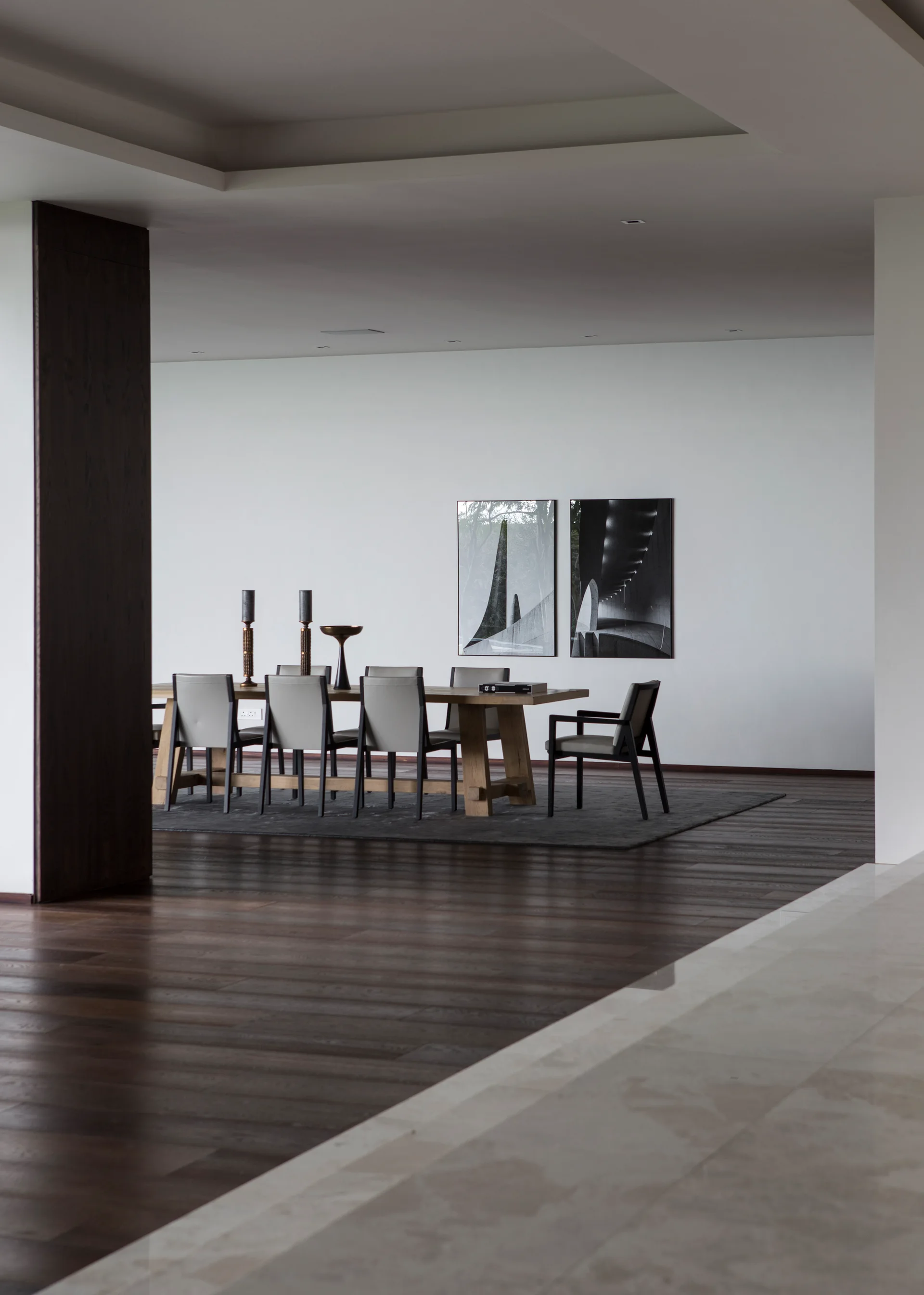 A minimalist dining room features a wooden table, upholstered grey chairs, dark wood flooring, and two black-and-white art prints on a white wall.