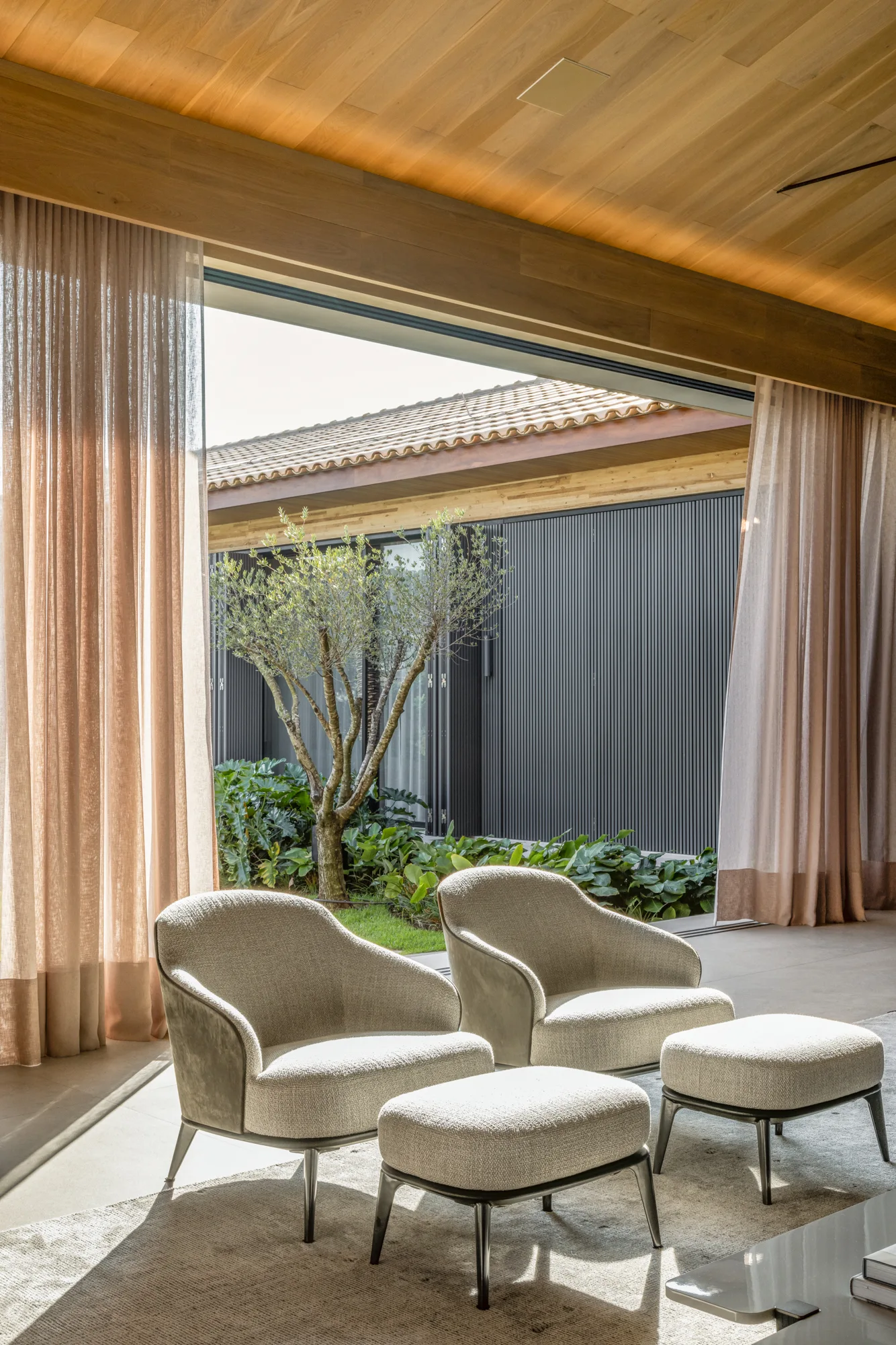 Two upholstered armchairs and matching ottomans sit on a rug under a wood ceiling, looking out to a courtyard with an olive tree.