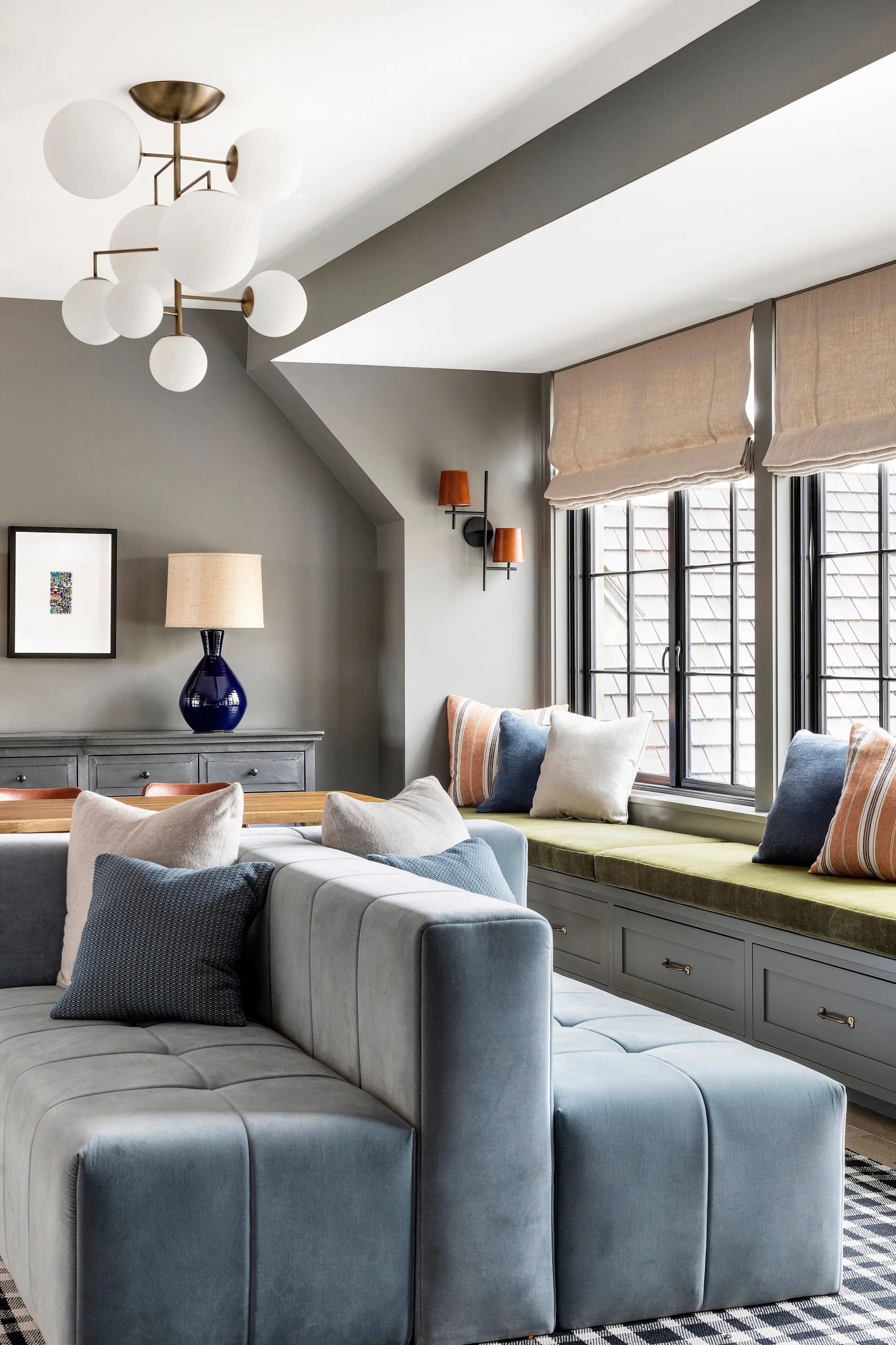 A grey living room features a blue modular sofa, a built-in window seat with an olive green cushion, and a multi-globe chandelier.