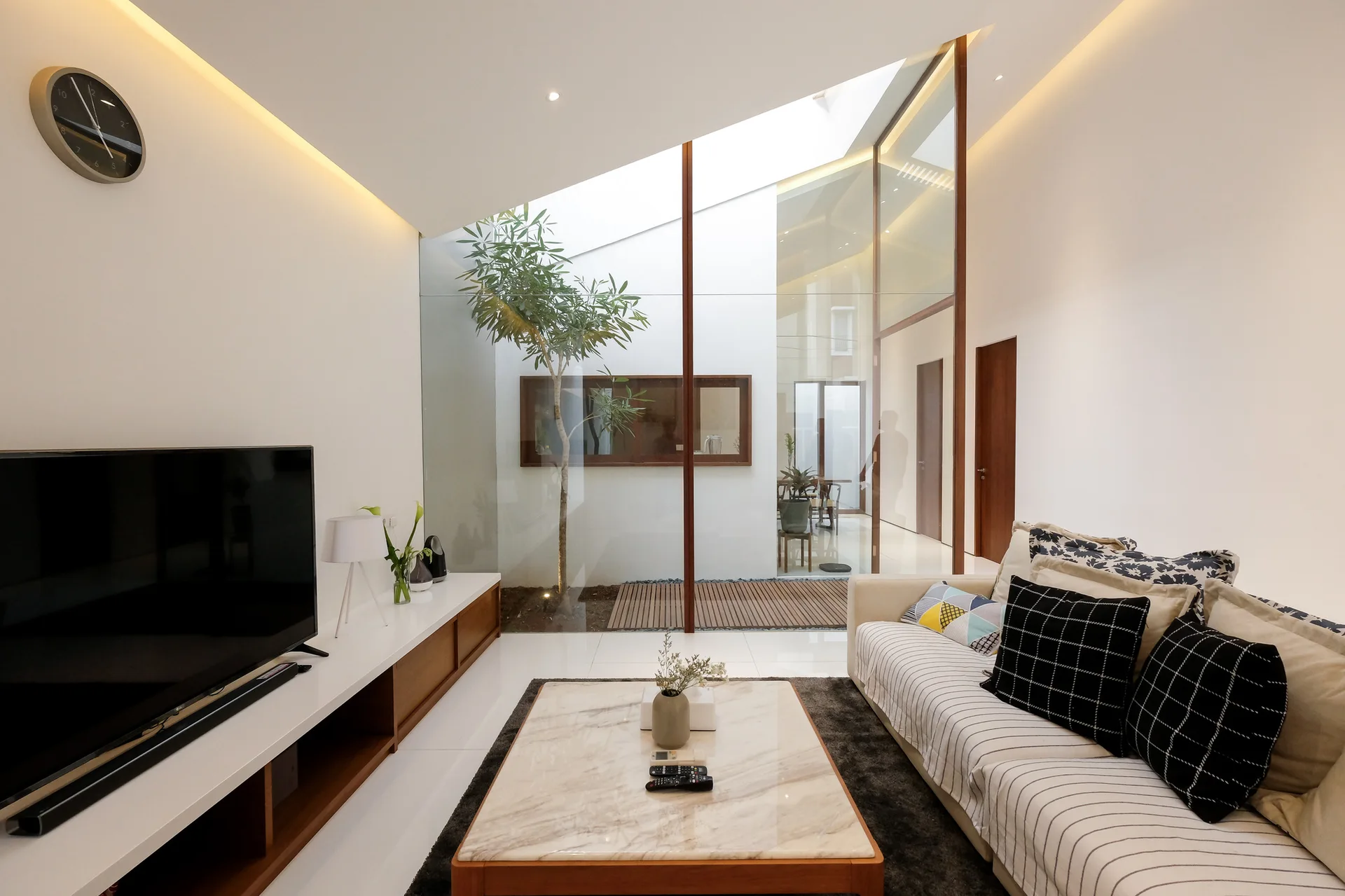 A modern living room features a striped sofa, a marble-topped coffee table, and a glass wall looking into an internal courtyard.