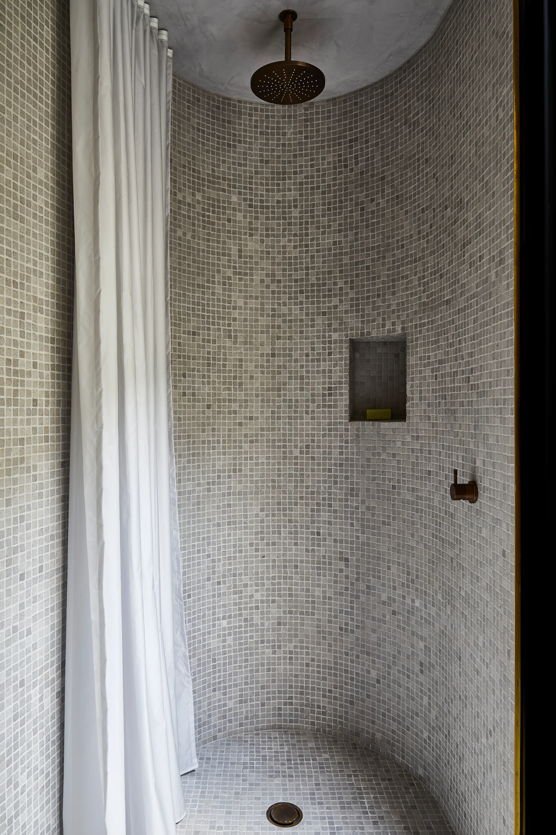 A curved bathroom shower stall features small gray mosaic tiling on the walls and floor, a white shower curtain, and dark bronze fixtures.