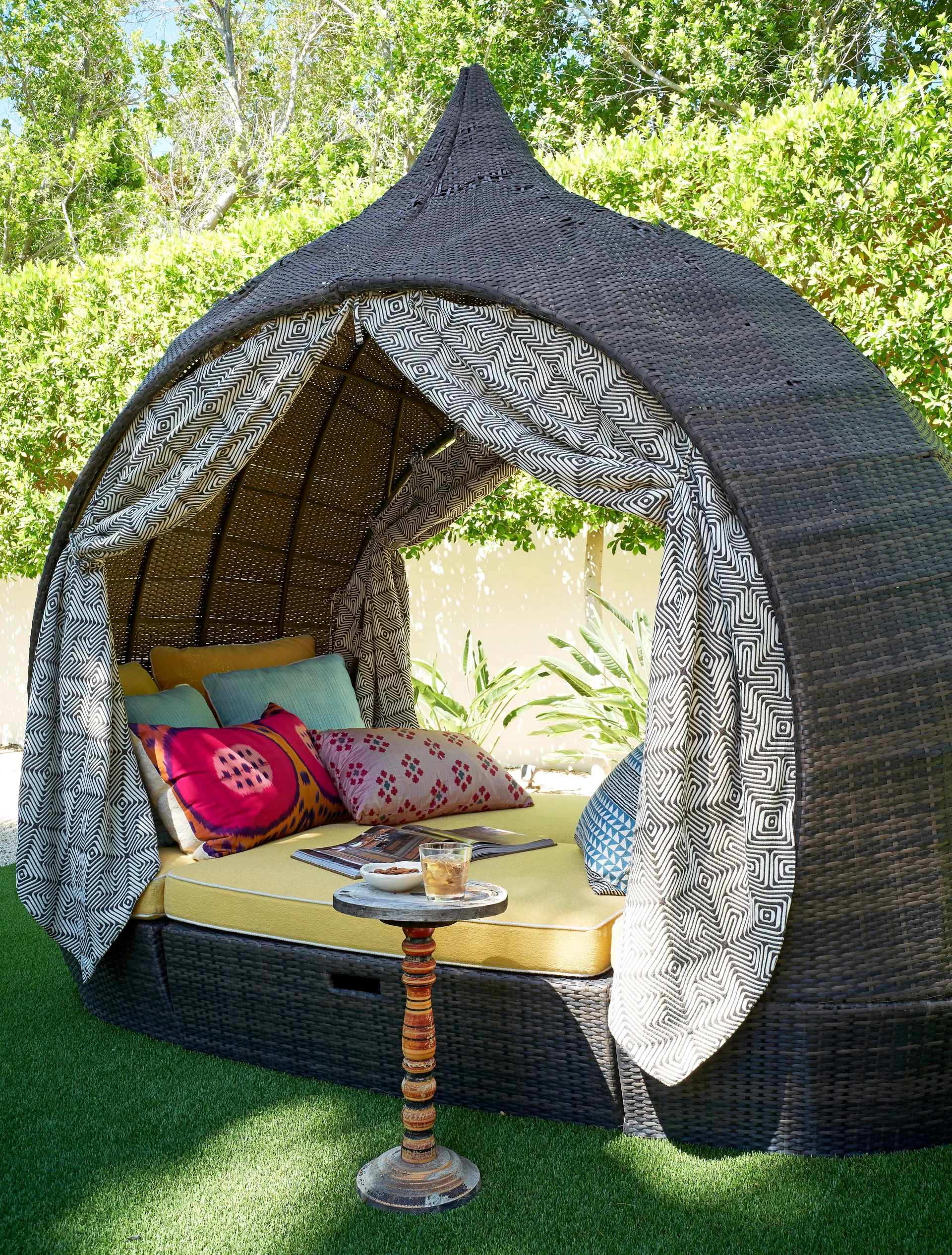 A dark woven wicker canopy daybed with yellow cushions and patterned curtains sits on a green lawn surrounded by garden foliage.