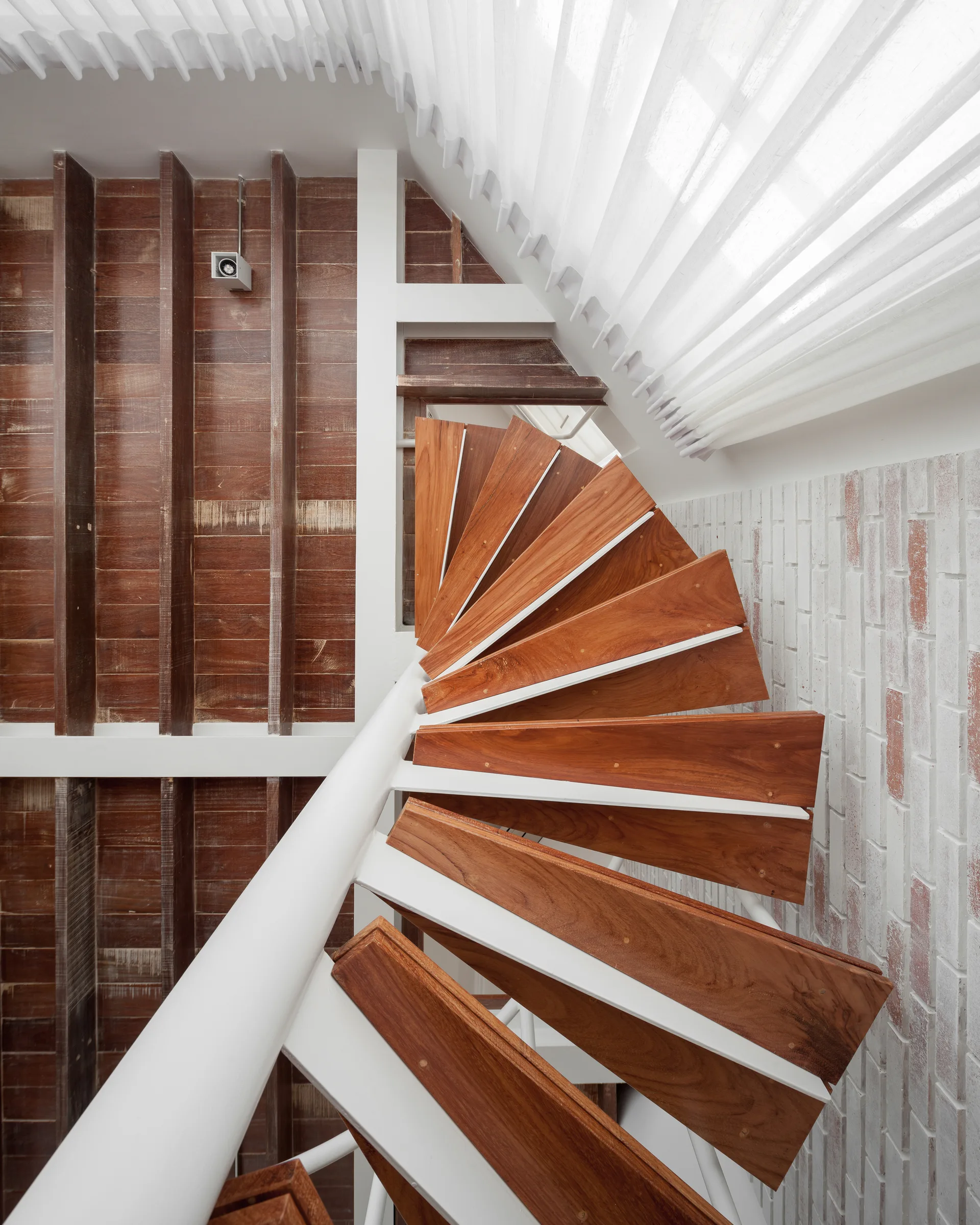 A top-down view of a modern spiral staircase with polished wood steps and a white handrail next to a white brick wall.