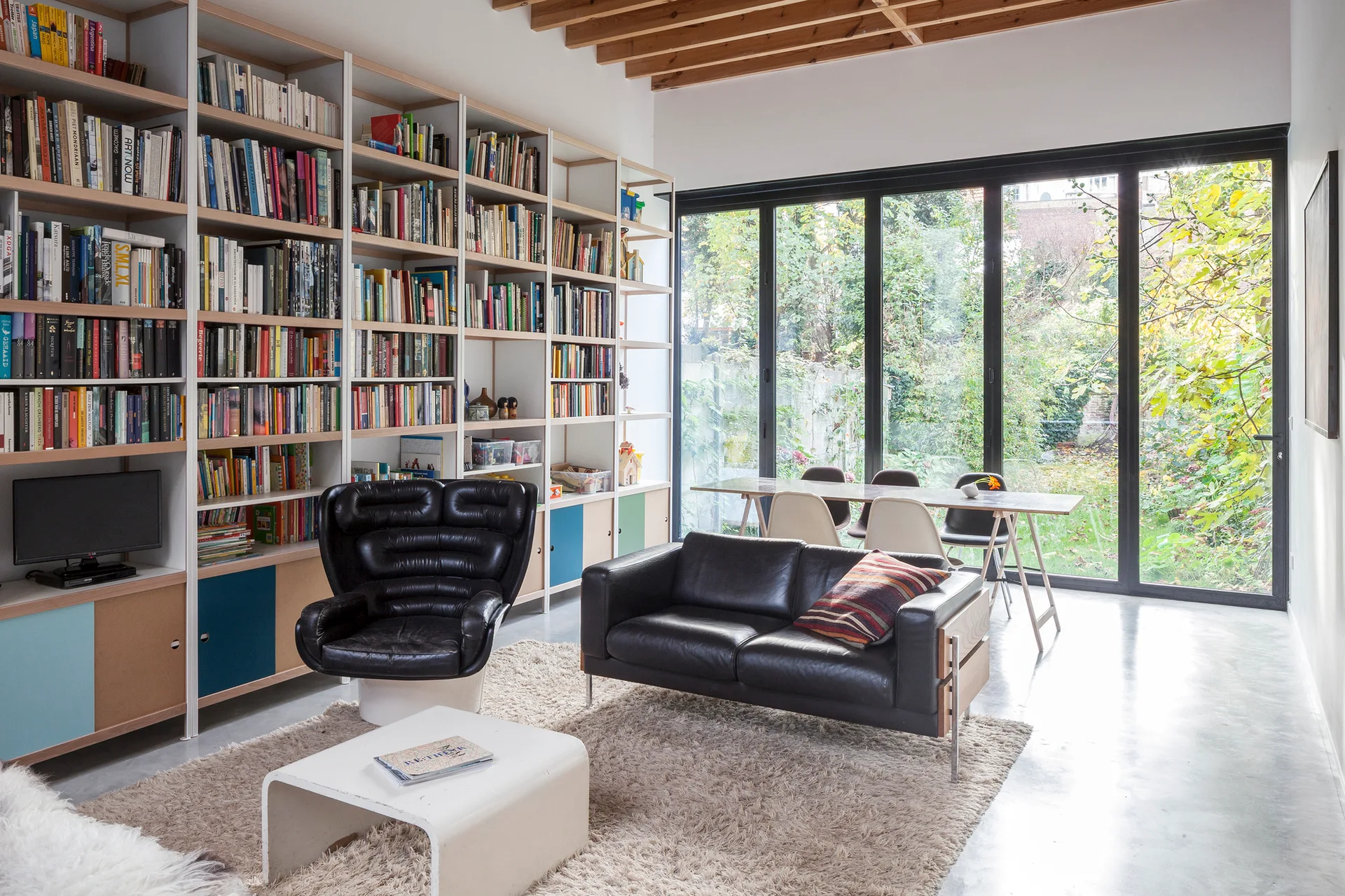 A bright living room features a massive floor-to-ceiling bookshelf, black leather furniture, and a glass wall opening to a green garden.