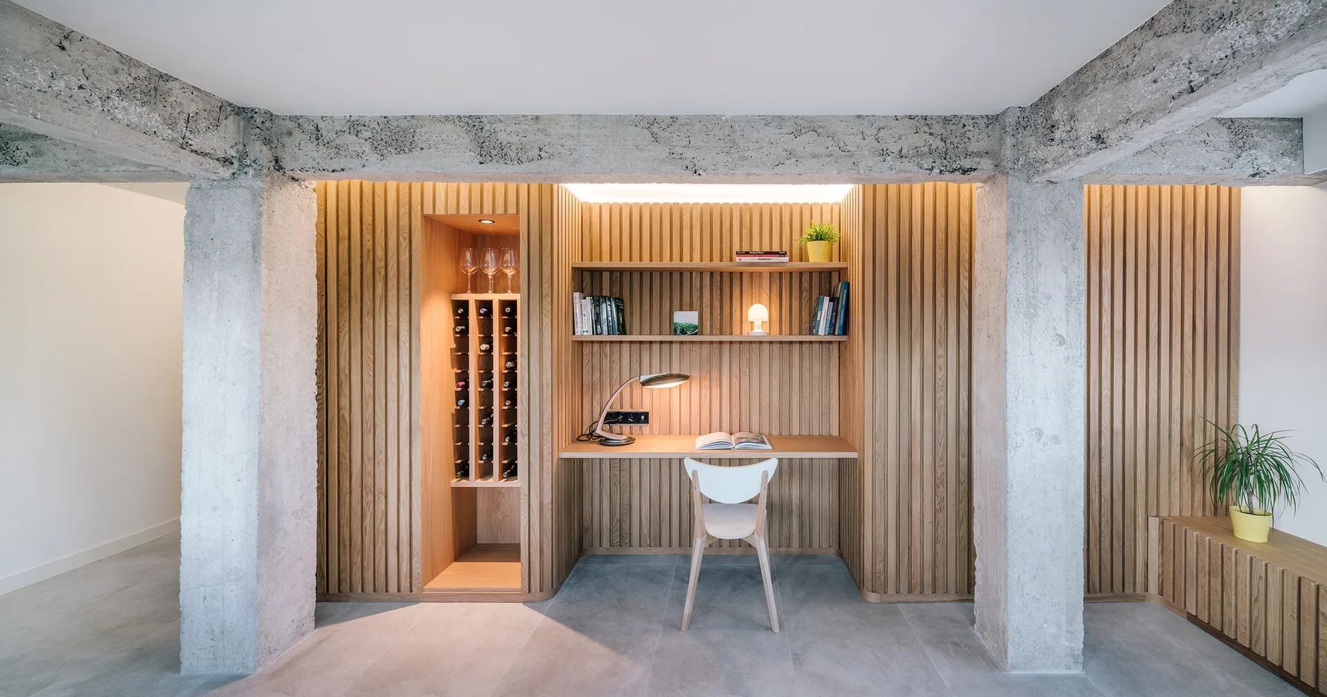A built-in wooden desk and wine rack are integrated into a vertical wood slat wall framed by two raw concrete columns.