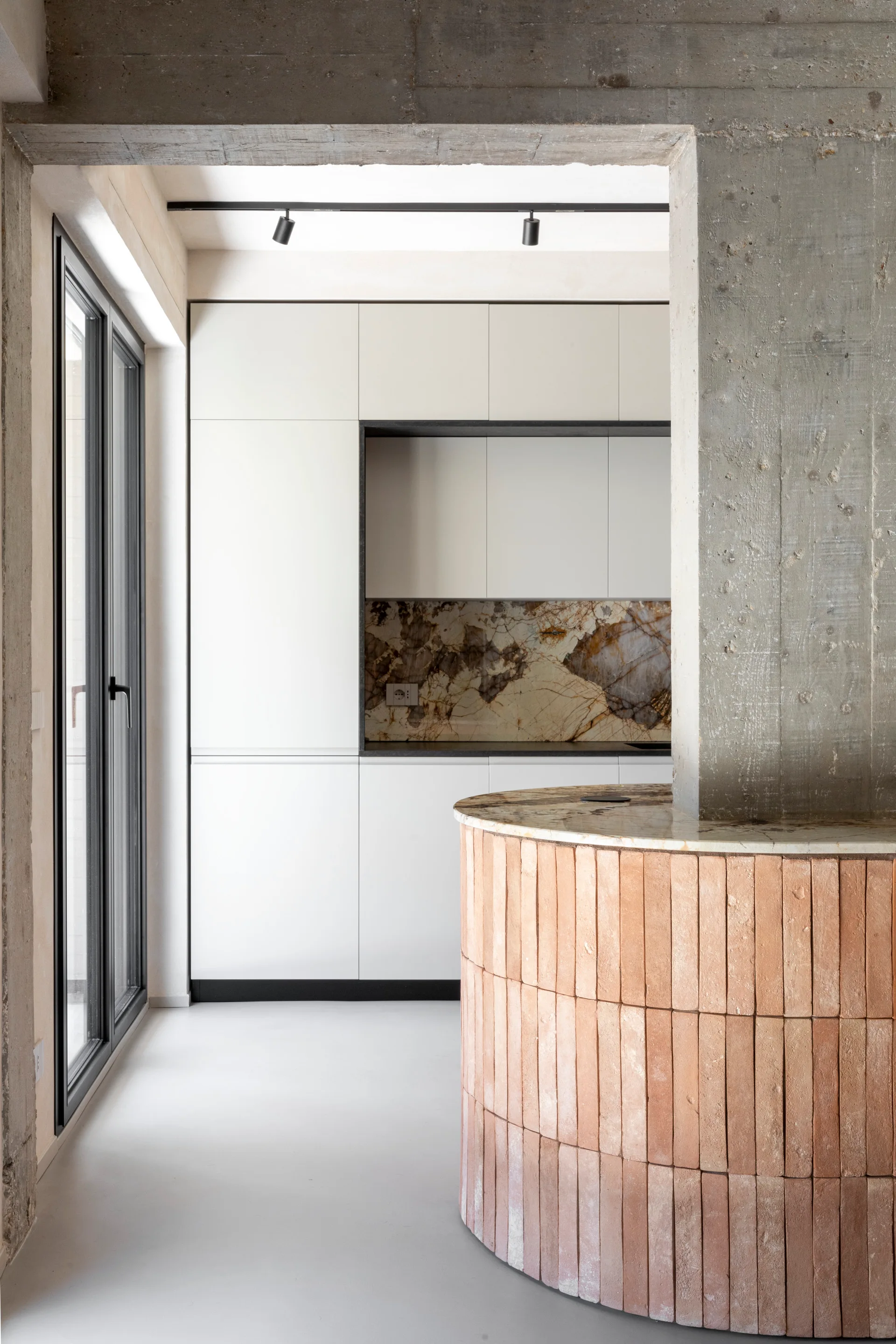 A kitchen featuring a curved island faced with terracotta bricks, minimalist white cabinetry, a veined stone backsplash, and a raw concrete column.