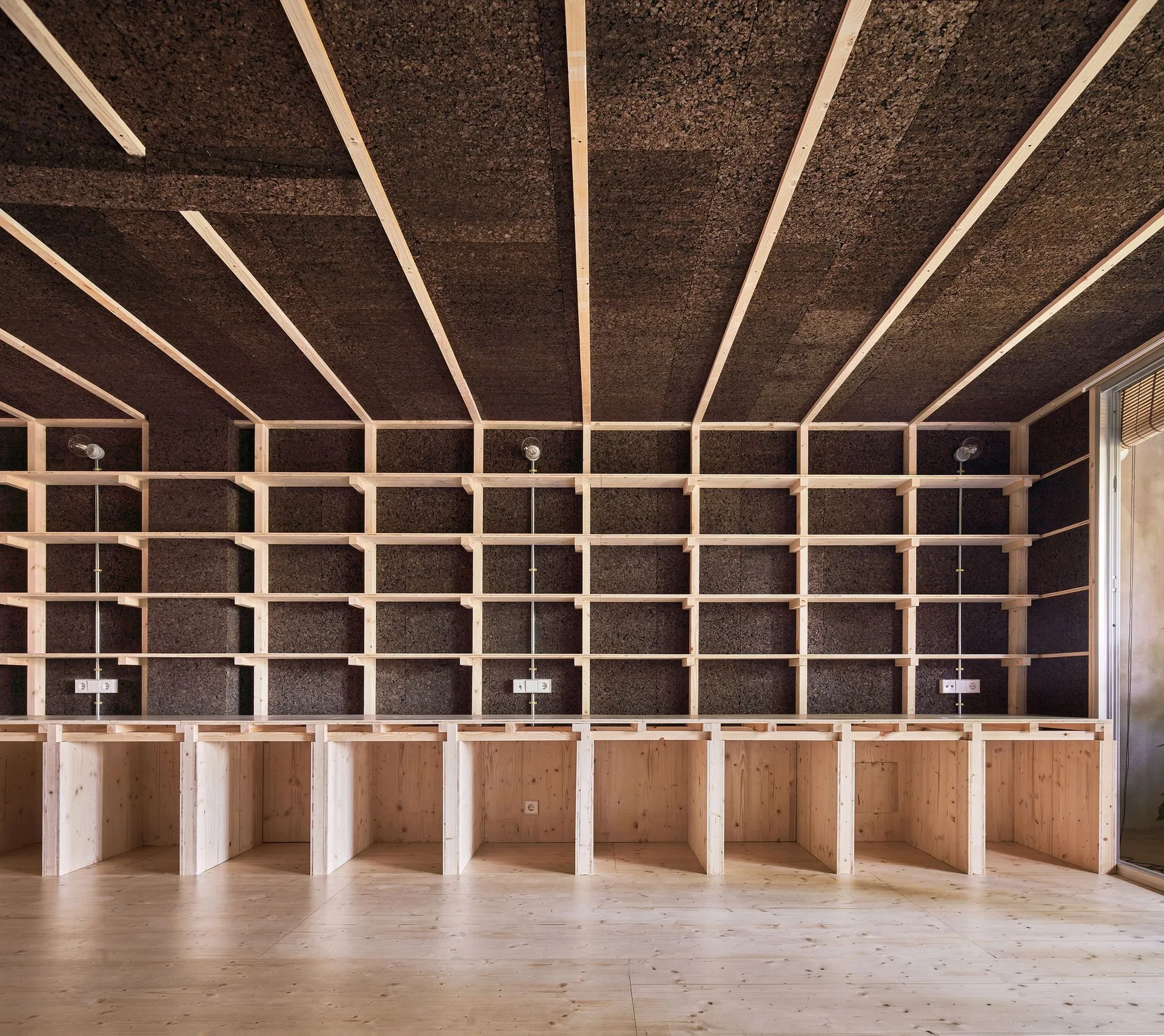 A modular light pine shelving unit covers a dark textured cork wall beneath a ceiling with matching timber rafters and exposed bulb light fixtures.