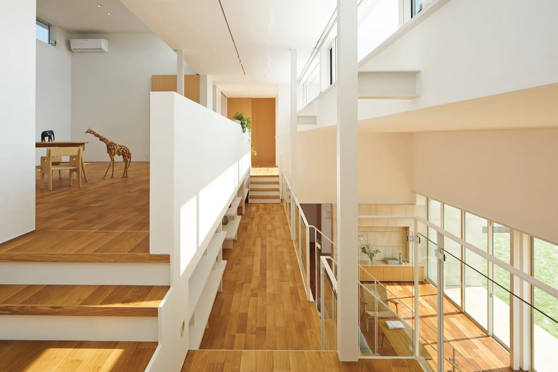 A minimalist split-level home interior featuring light wood flooring, white walls, built-in shelves, and glass railings overlooking a lower level.