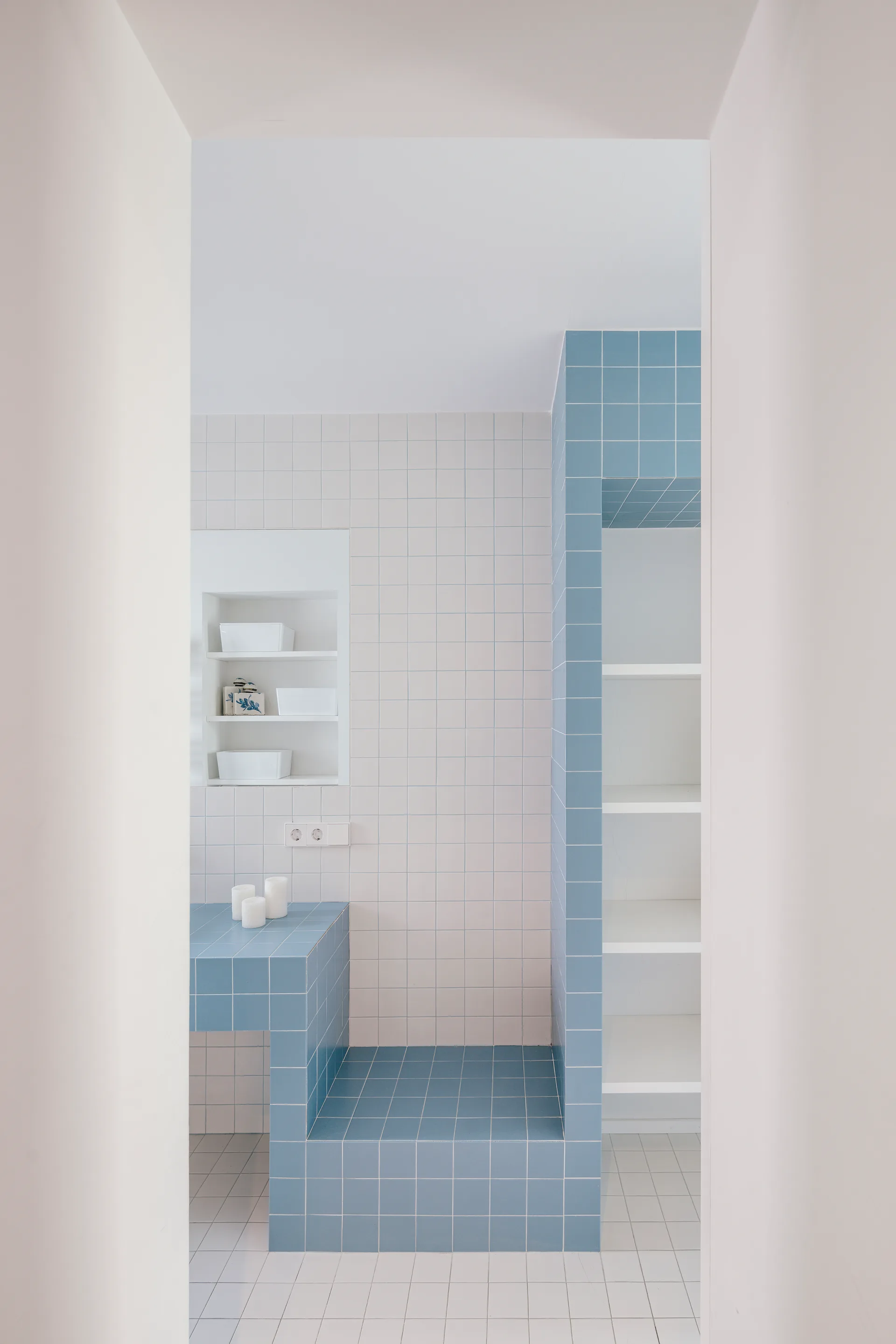 A minimalist bathroom featuring white grid walls, a recessed wall niche, and sculptural built-in shelving constructed with light blue tiles.