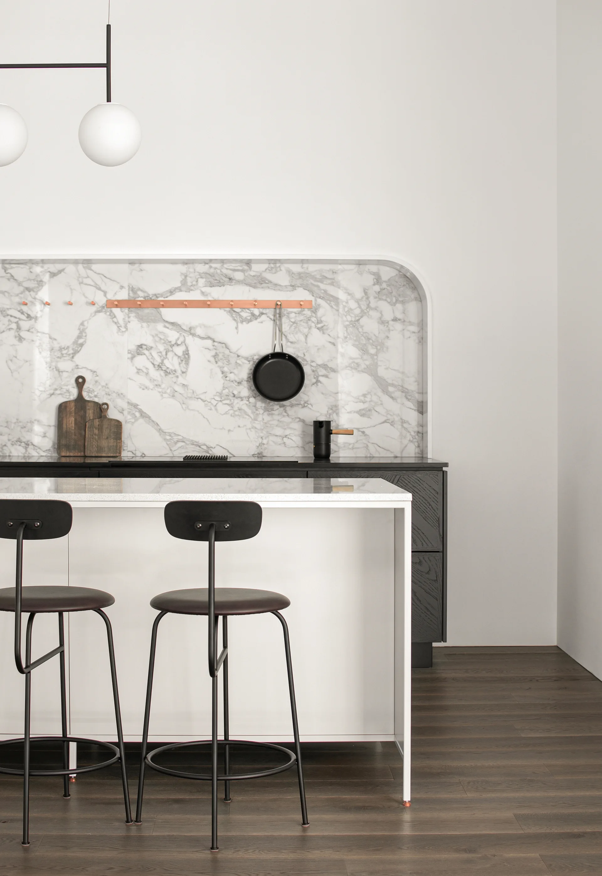 A modern kitchen featuring a white island with two black stools in front of an arched marble backsplash with a copper hanging rail.