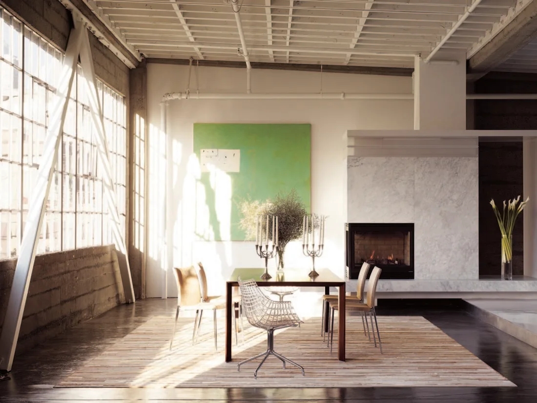 An industrial loft dining room features a dark wood table with mixed chairs, a large marble fireplace, and a massive multi-pane window.
