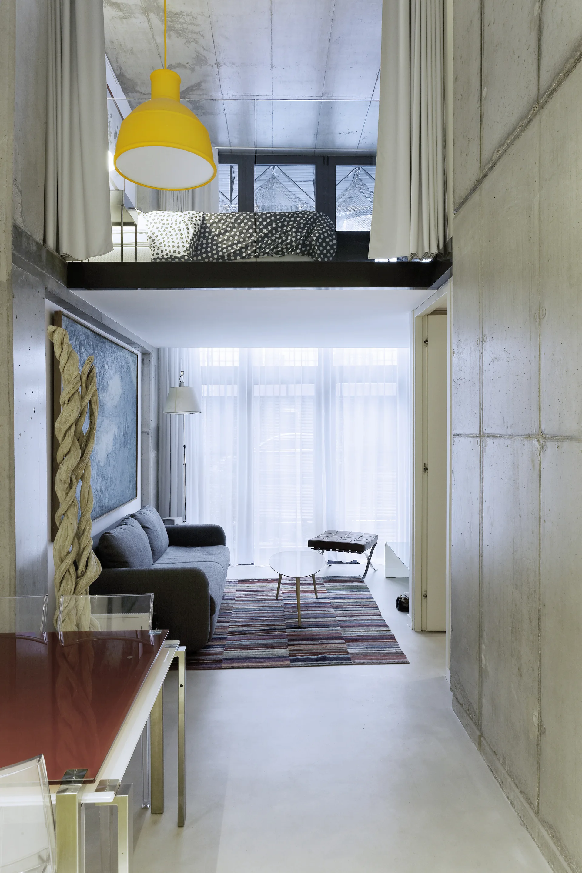 A modern industrial loft features a gray sofa, a striped rug, exposed concrete walls, and a mezzanine bedroom above with a yellow pendant light.