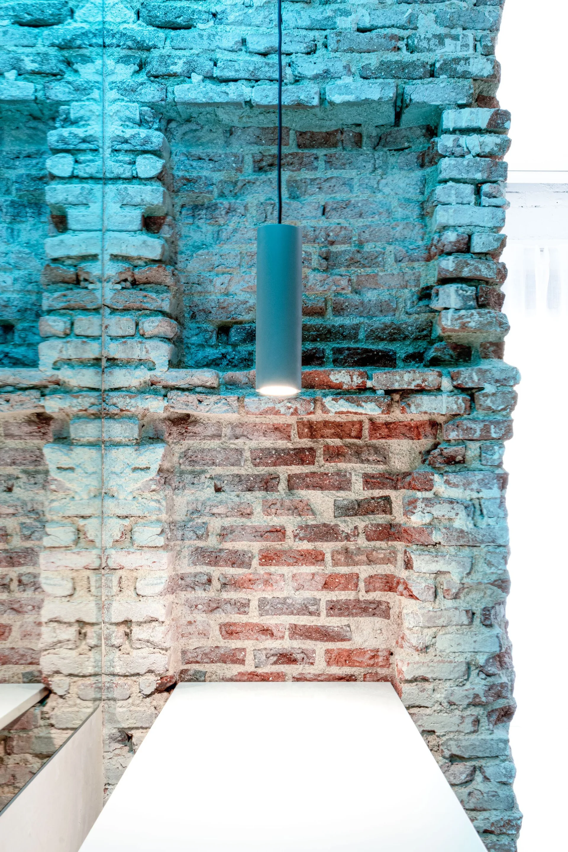 A vertical mirror reflects an exposed red brick wall illuminated by a cylindrical grey pendant light hanging over a white tabletop.