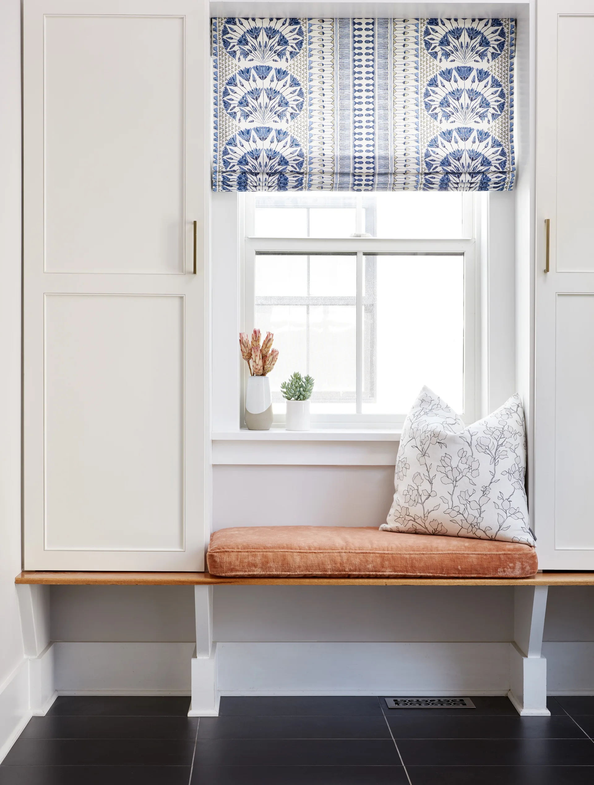 A built-in window seat with an orange velvet cushion is flanked by tall white cabinets under a blue and white patterned Roman shade.