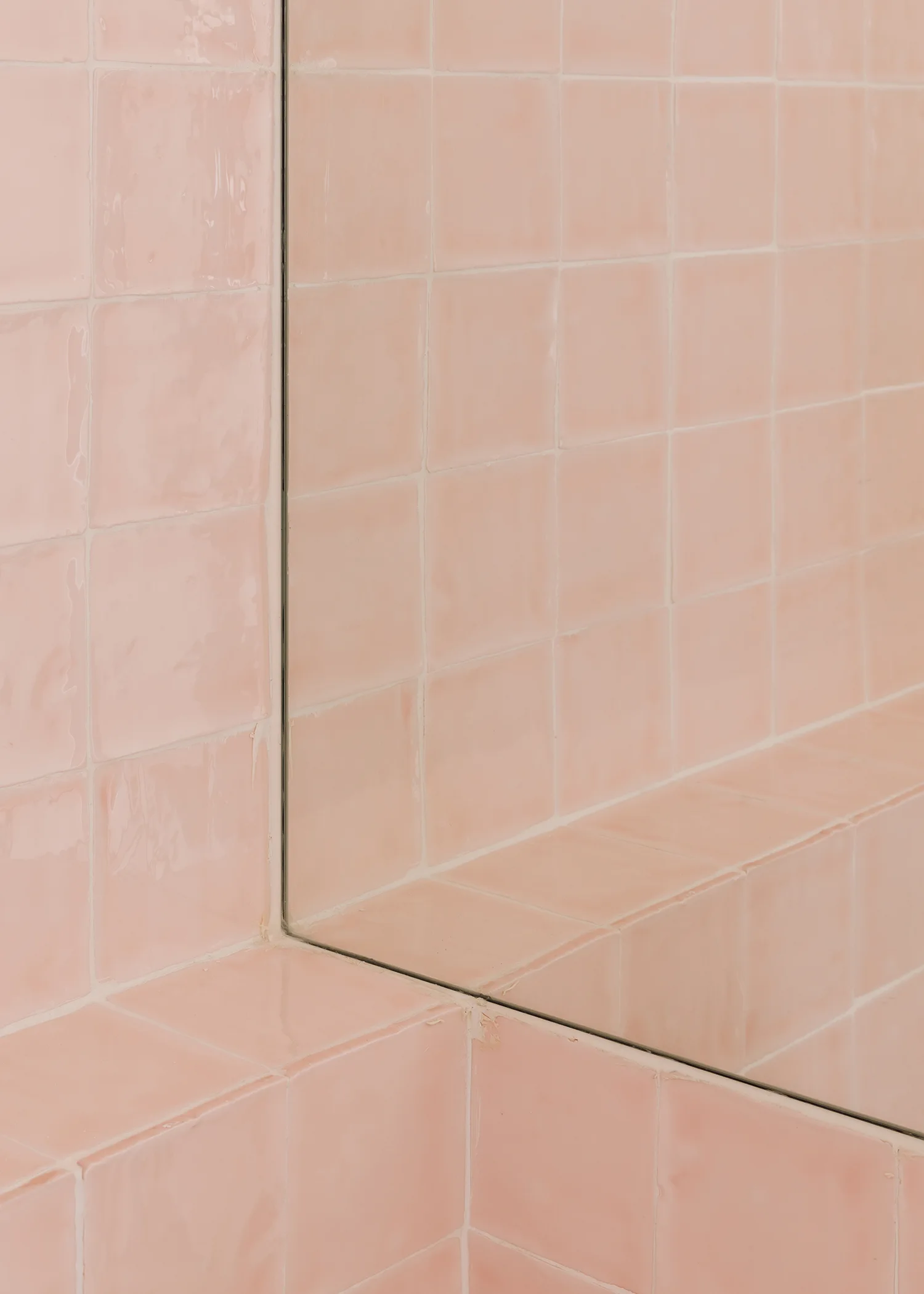 A close-up corner view of glossy blush pink square ceramic tiles on a bathroom wall meeting a frameless inset mirror.