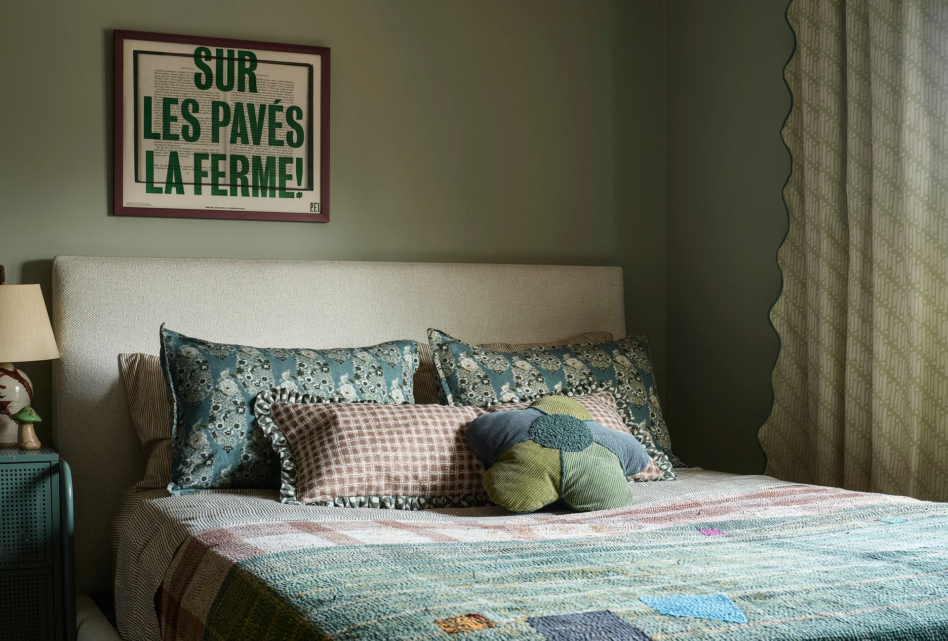 A cozy bedroom features a neutral upholstered headboard against a sage green wall with an eclectic mix of patterned throw pillows.