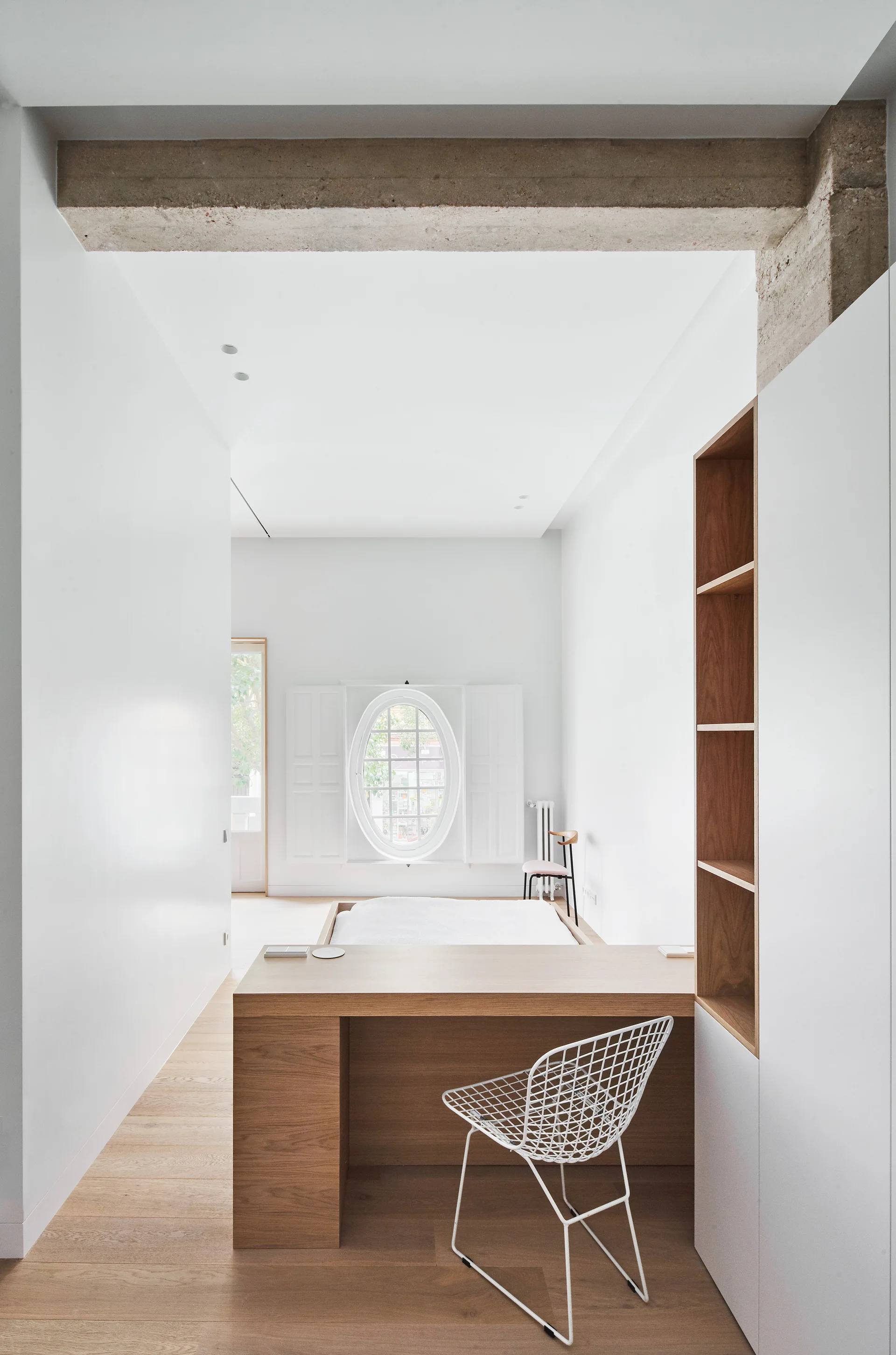A minimalist white bedroom featuring an integrated wood desk, a low platform bed, an oval window with shutters, and an exposed concrete ceiling beam.