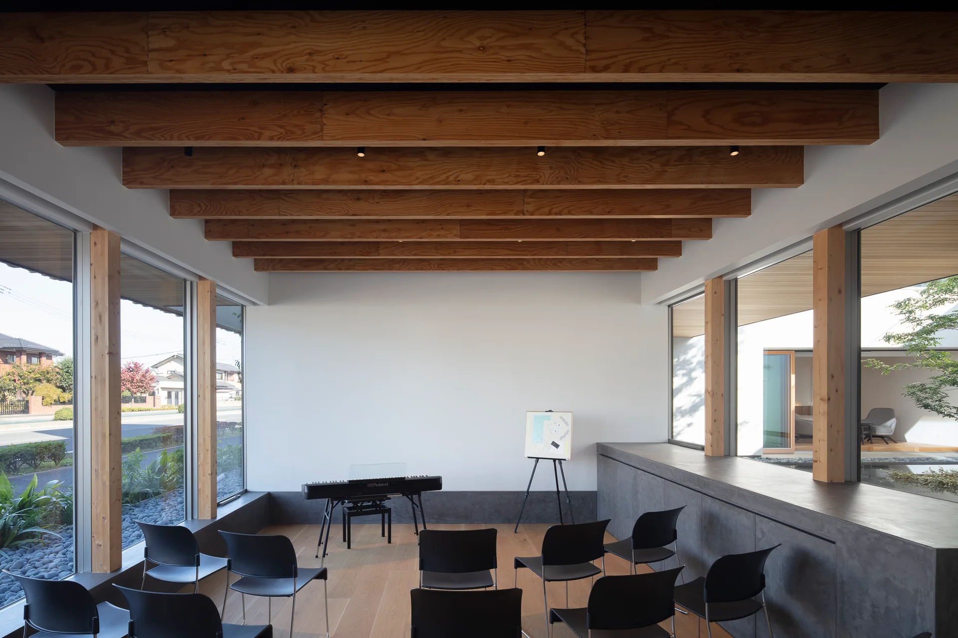A minimalist studio features rows of black chairs facing a digital piano under a high ceiling with prominent exposed wood beams.