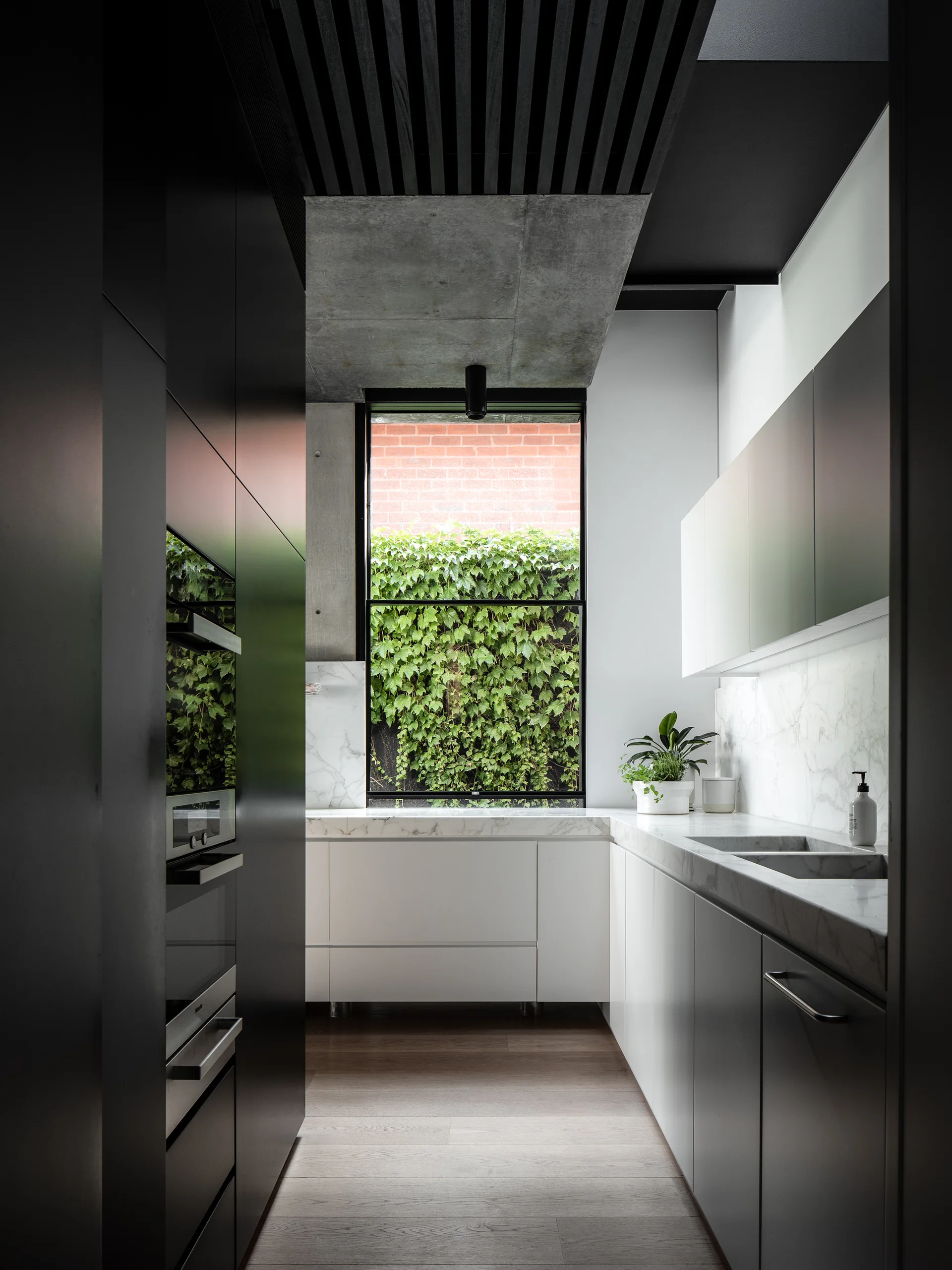 A narrow modern kitchen features matte black and white cabinets, marble countertops, and a large window overlooking a lush ivy wall.
