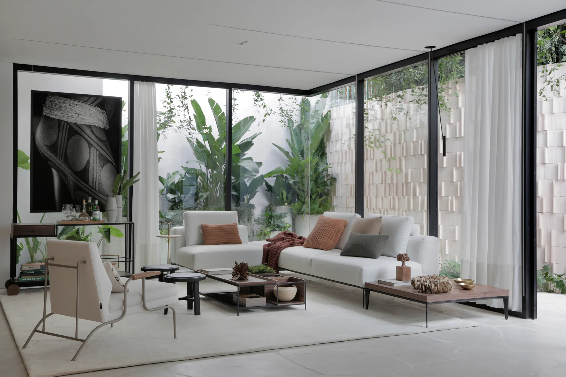 A modern minimalist living room features white modular sofas and low wooden coffee tables set against floor-to-ceiling glass walls looking onto a garden.