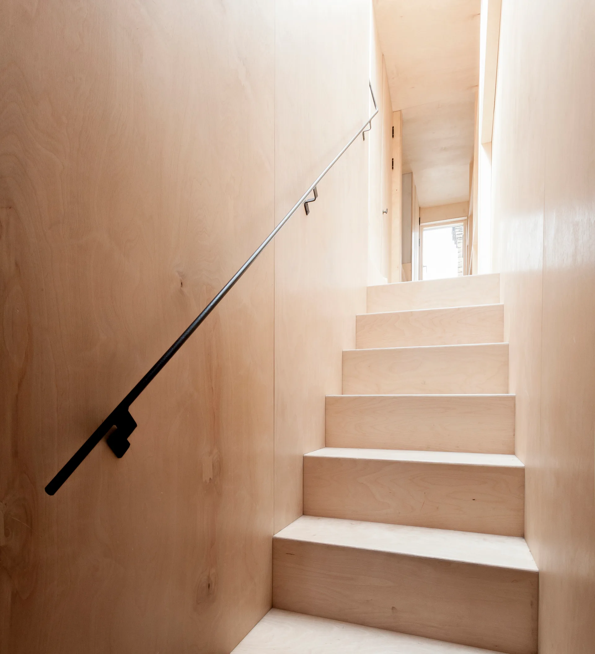 A minimalist staircase and surrounding walls constructed entirely from light-toned plywood, featuring a slim black metal handrail.
