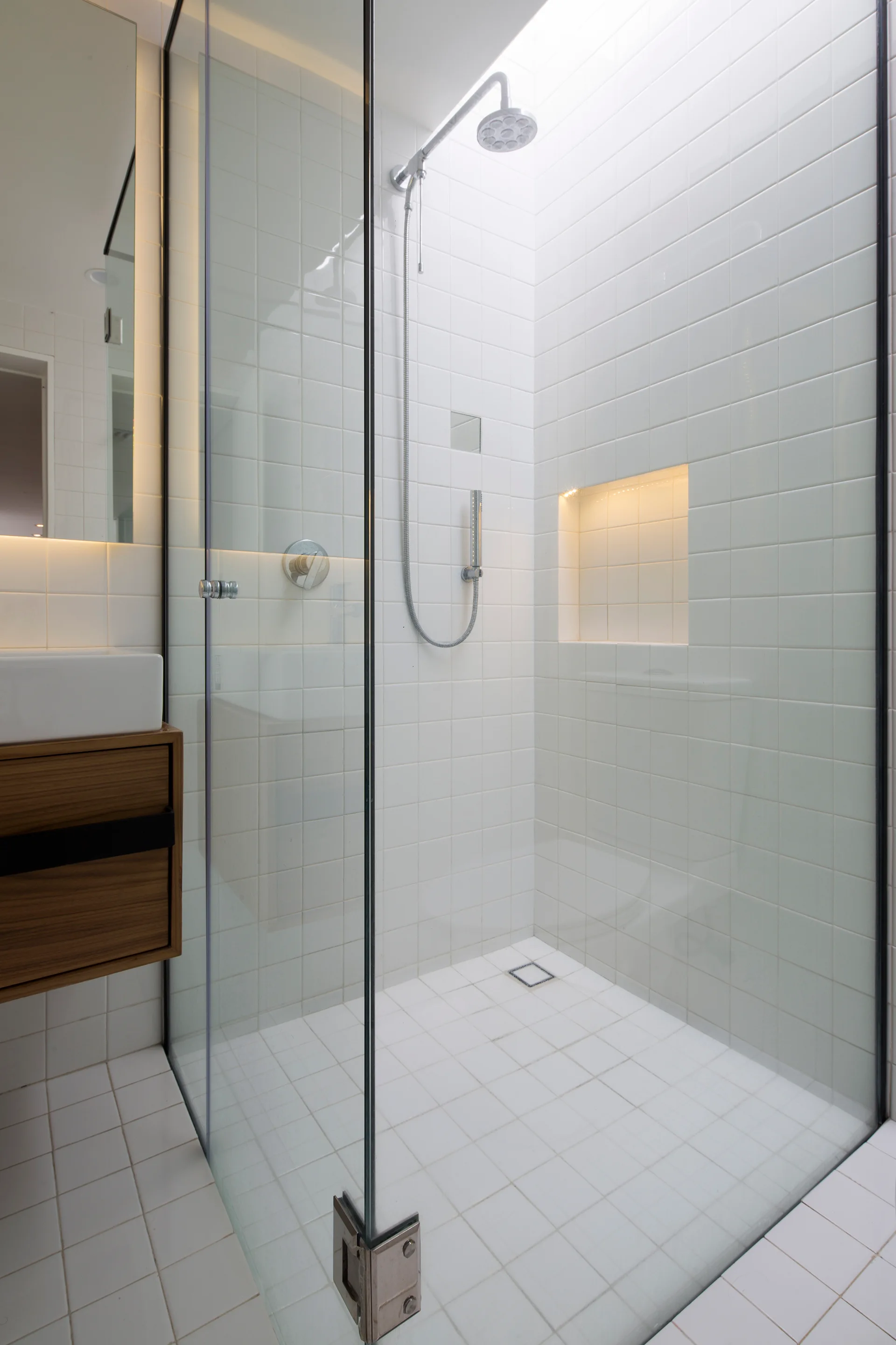 A modern bathroom features a glass shower enclosure with white square tiled walls and floor, a chrome shower head, and an illuminated recessed niche.