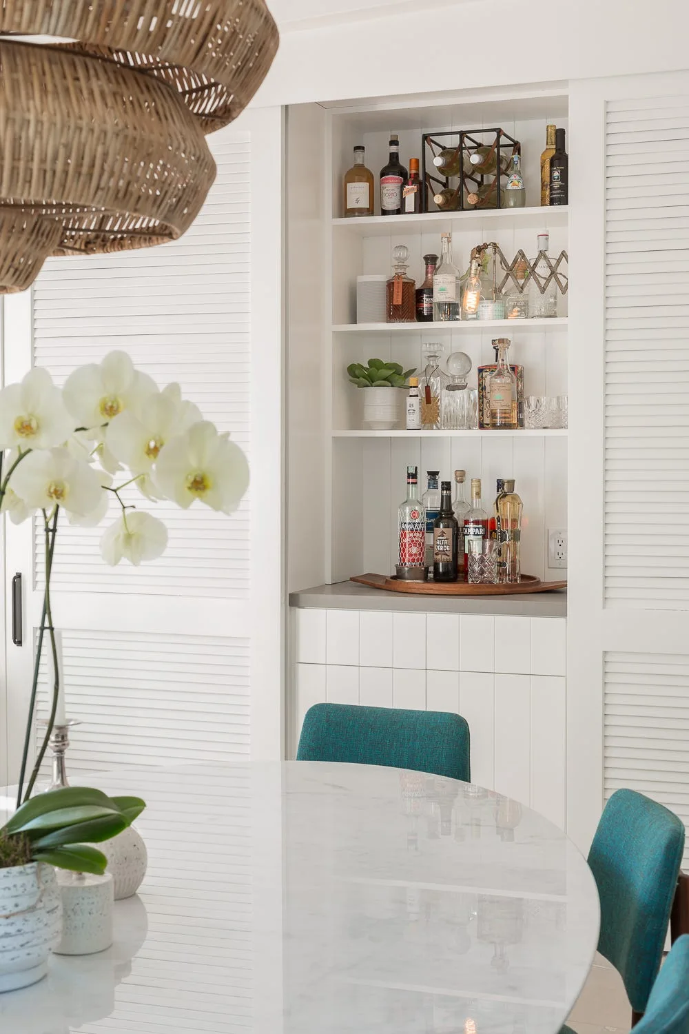 A bright dining area features a marble table and teal chairs in front of a white recessed home bar cabinet with sliding louvered doors.