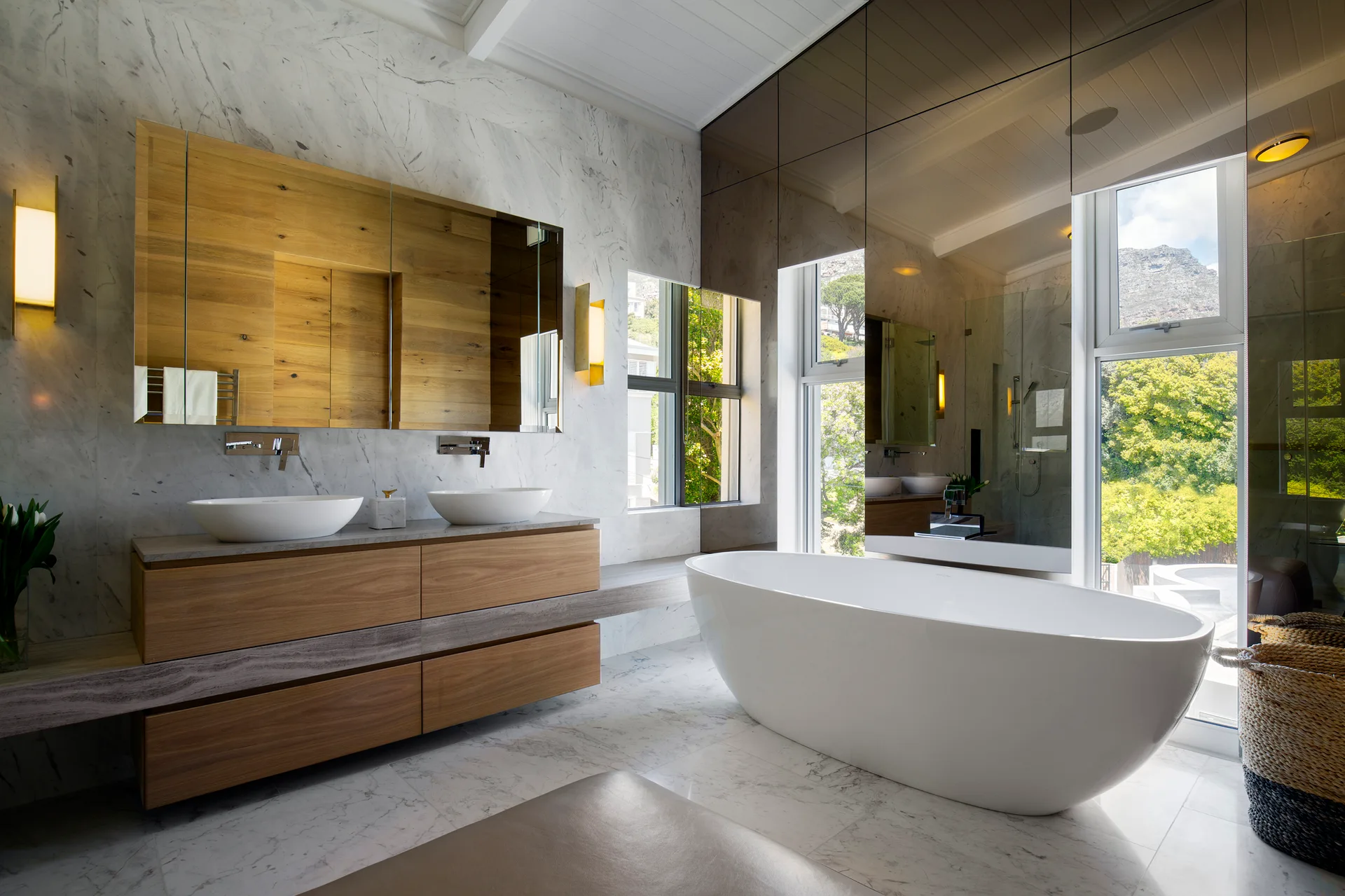 A modern bathroom features a white freestanding bathtub alongside a floating wood vanity with double sinks, surrounded by veined marble walls.