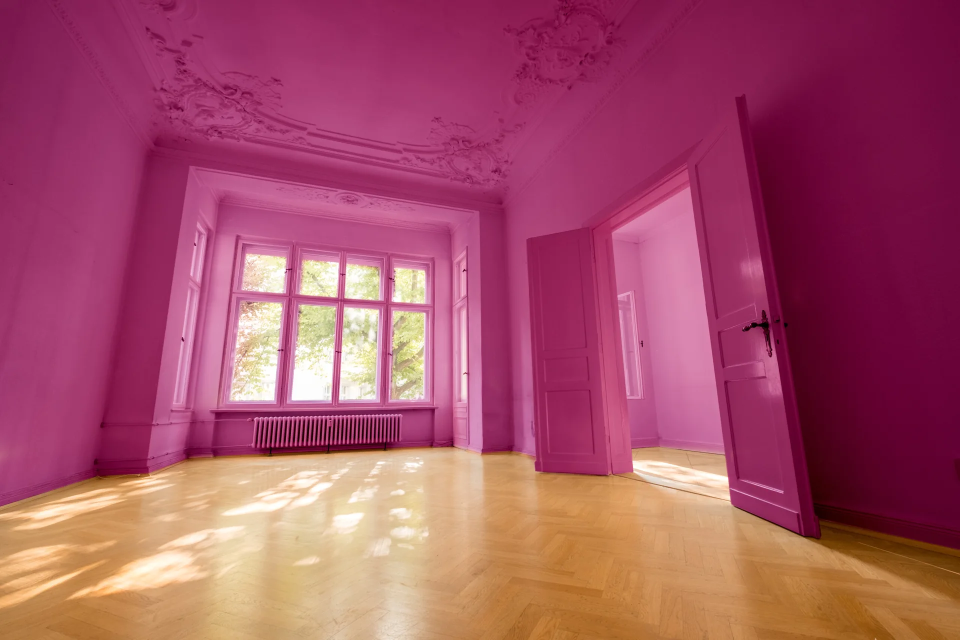 An empty neoclassical room with walls, ceiling, and doors painted in solid magenta above light-toned wood herringbone floors.