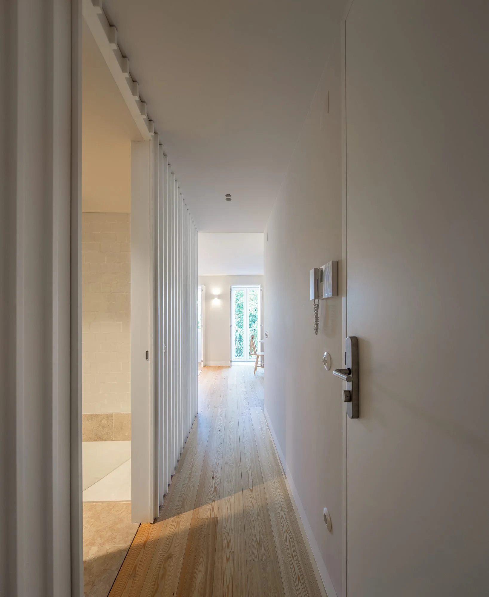 A minimalist hallway with clean white walls, light wood plank flooring, and a white slatted partition screen leading to an adjacent room.