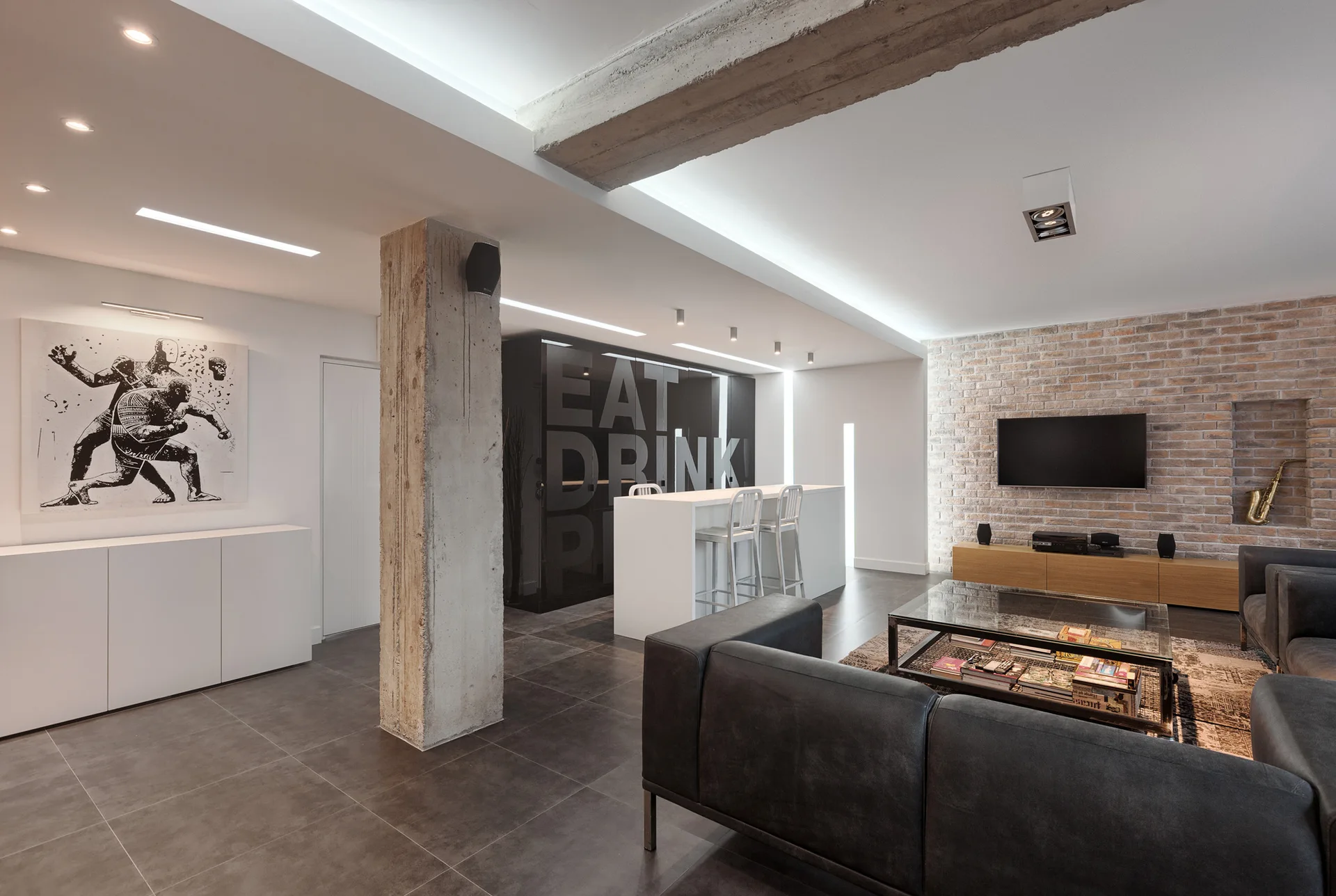 An industrial open-plan living room featuring a grey sectional sofa, an exposed brick wall with a television, and a raw concrete structural column.