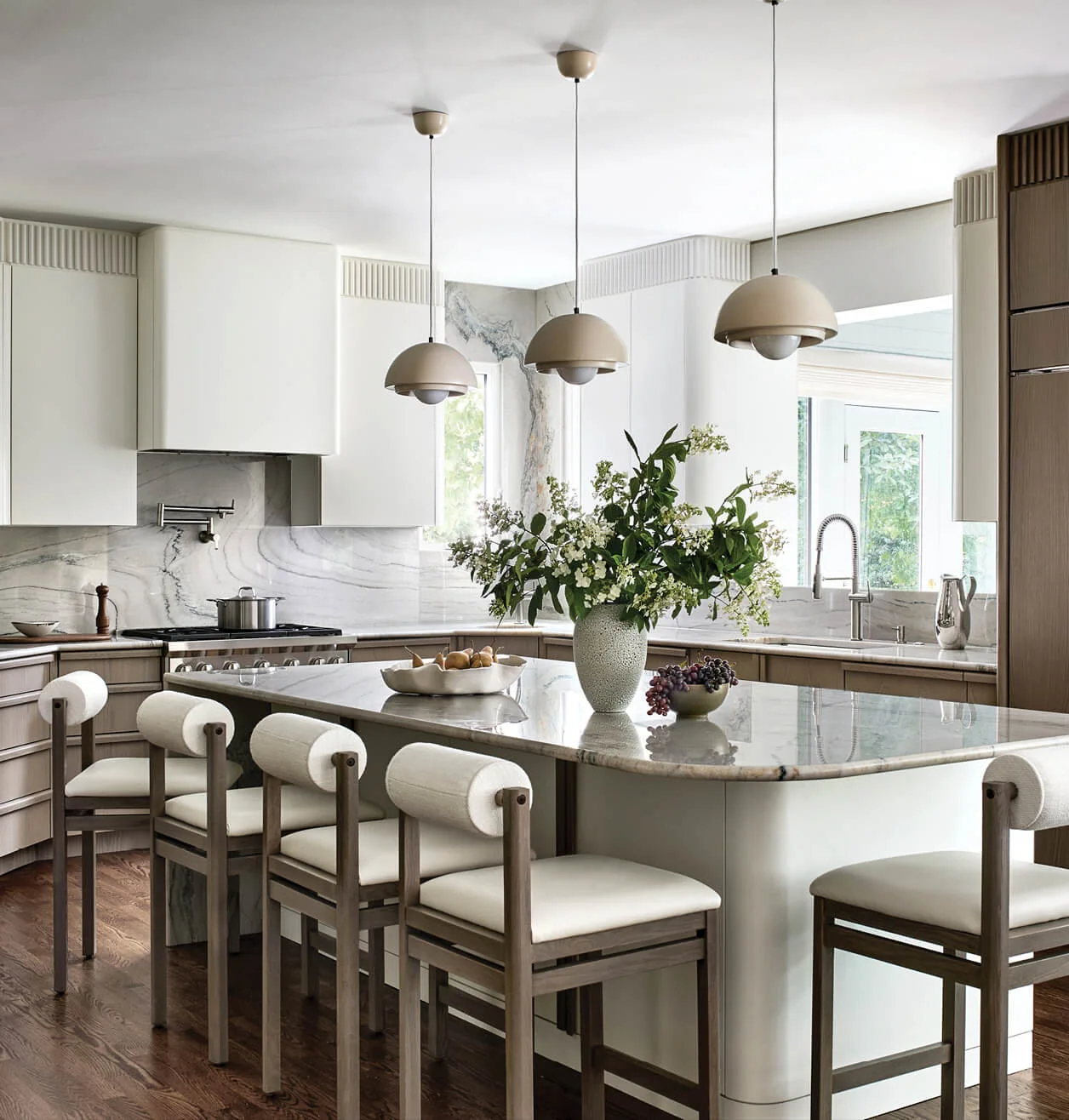A bright kitchen features a curved stone-topped island with wood bar stools, marble backsplash, and three taupe dome pendant lights.