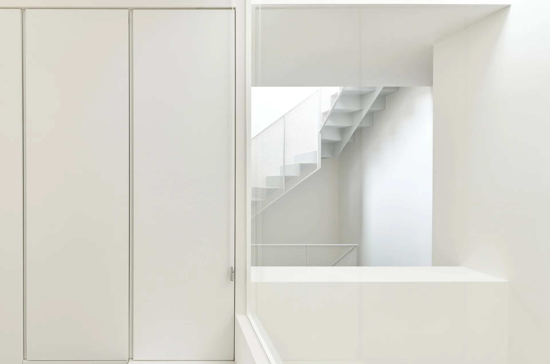 An all-white minimalist interior with a glass partition looking into a staircase featuring a white metal mesh railing.