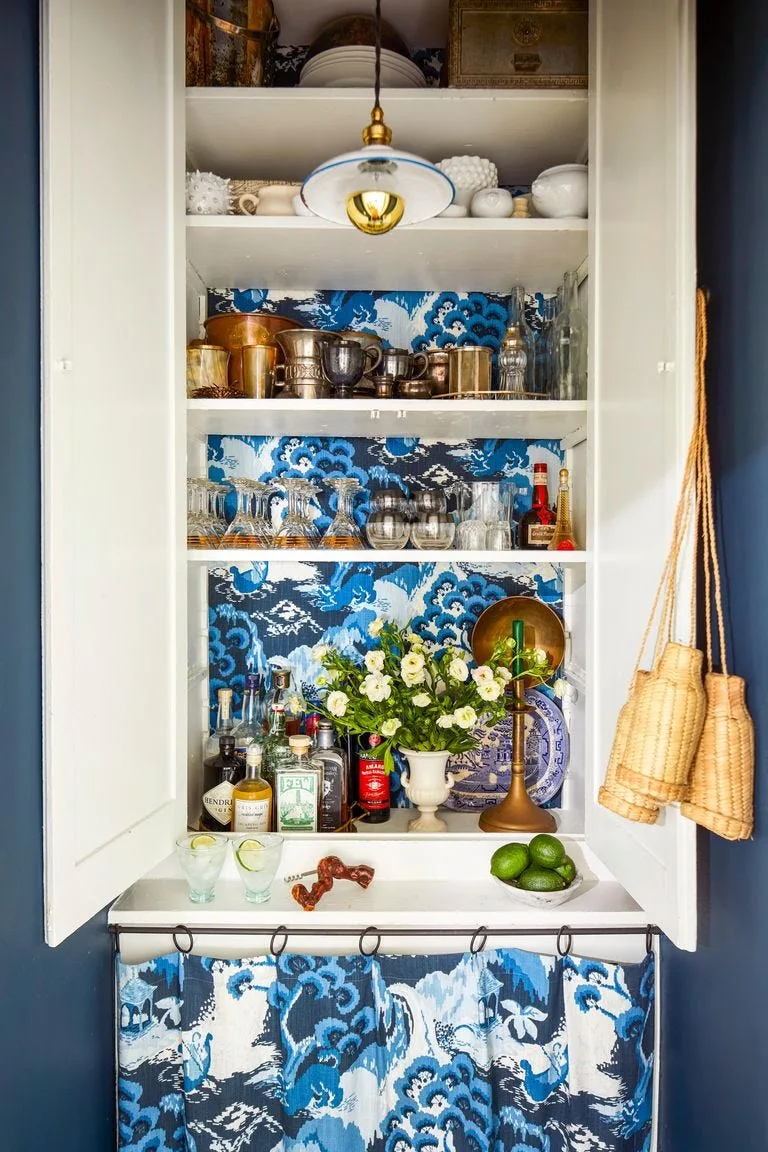 A white built-in bar cabinet features shelves lined with blue and white patterned wallpaper, stocked with liquor bottles, glassware, and a pendant light.